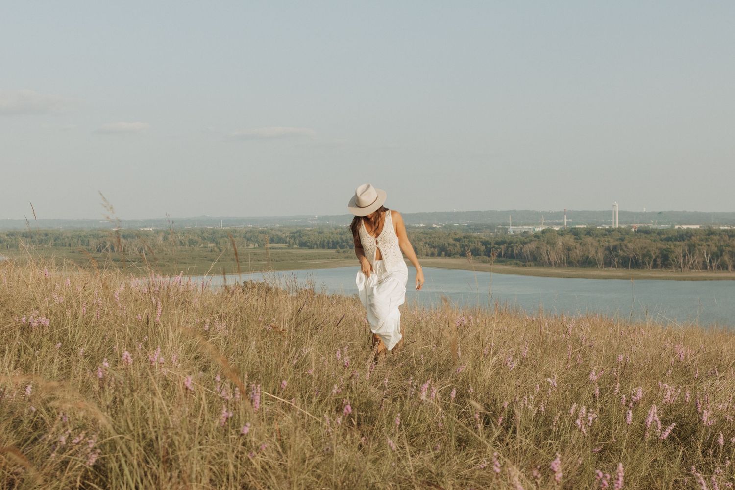 A person in a white dress walks through a scenic field of tall grass near a lake on a sunny day.