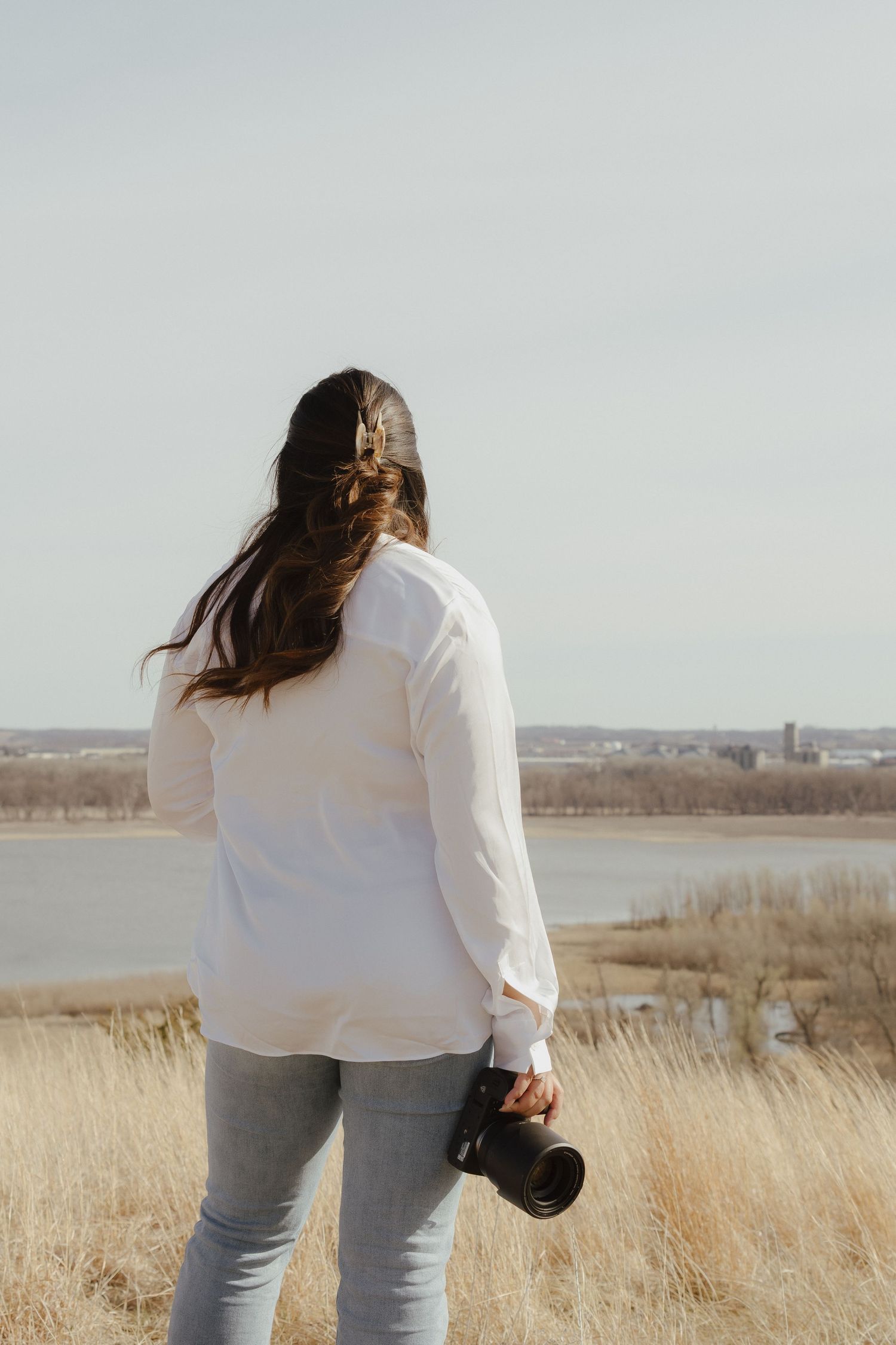 A person in a white sweater and jeans stands overlooking a lake with tall grass in the foreground, holding a camera.