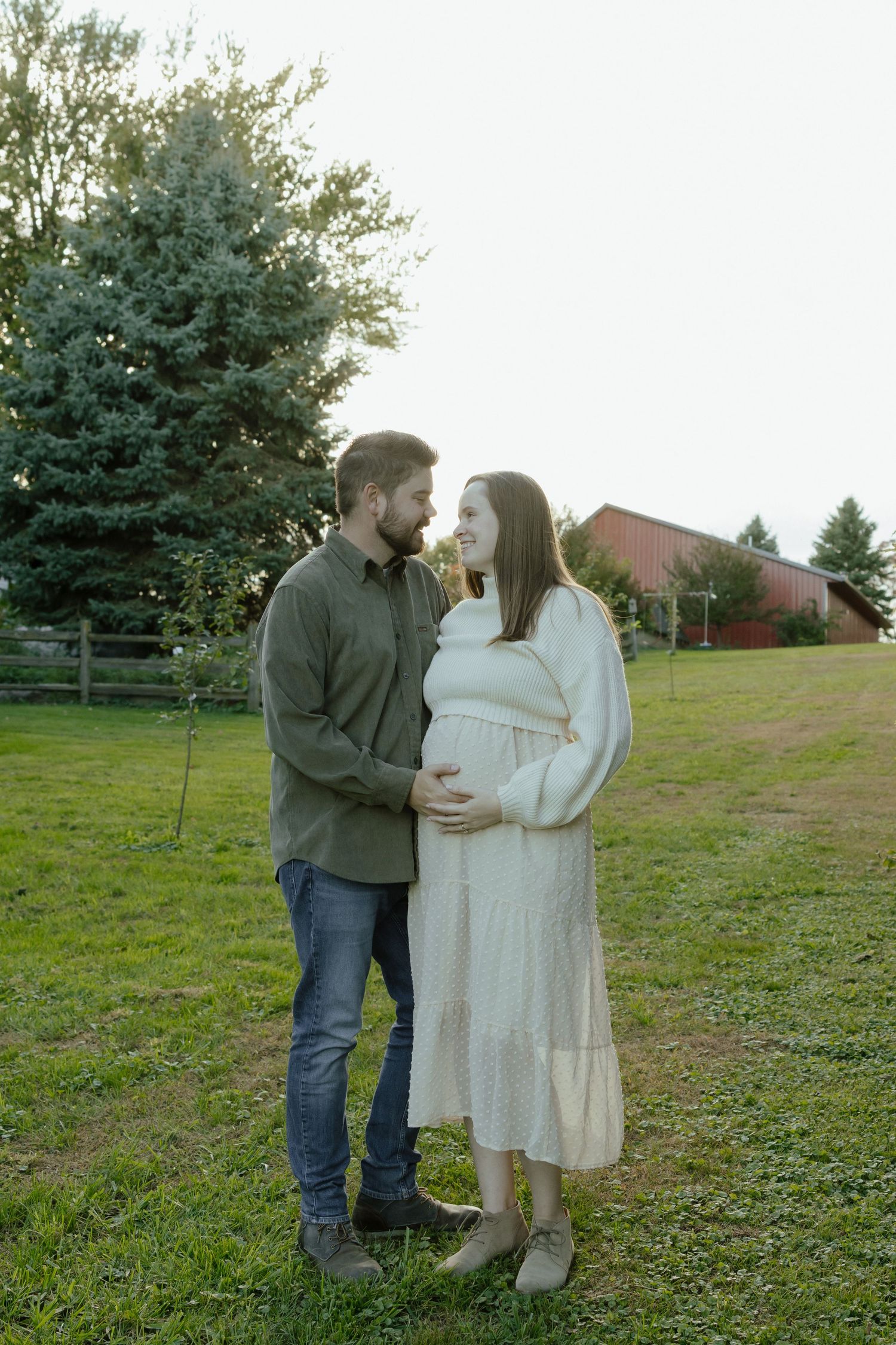 A couple embraces on a grassy hillside with evergreen trees and buildings in the background.
