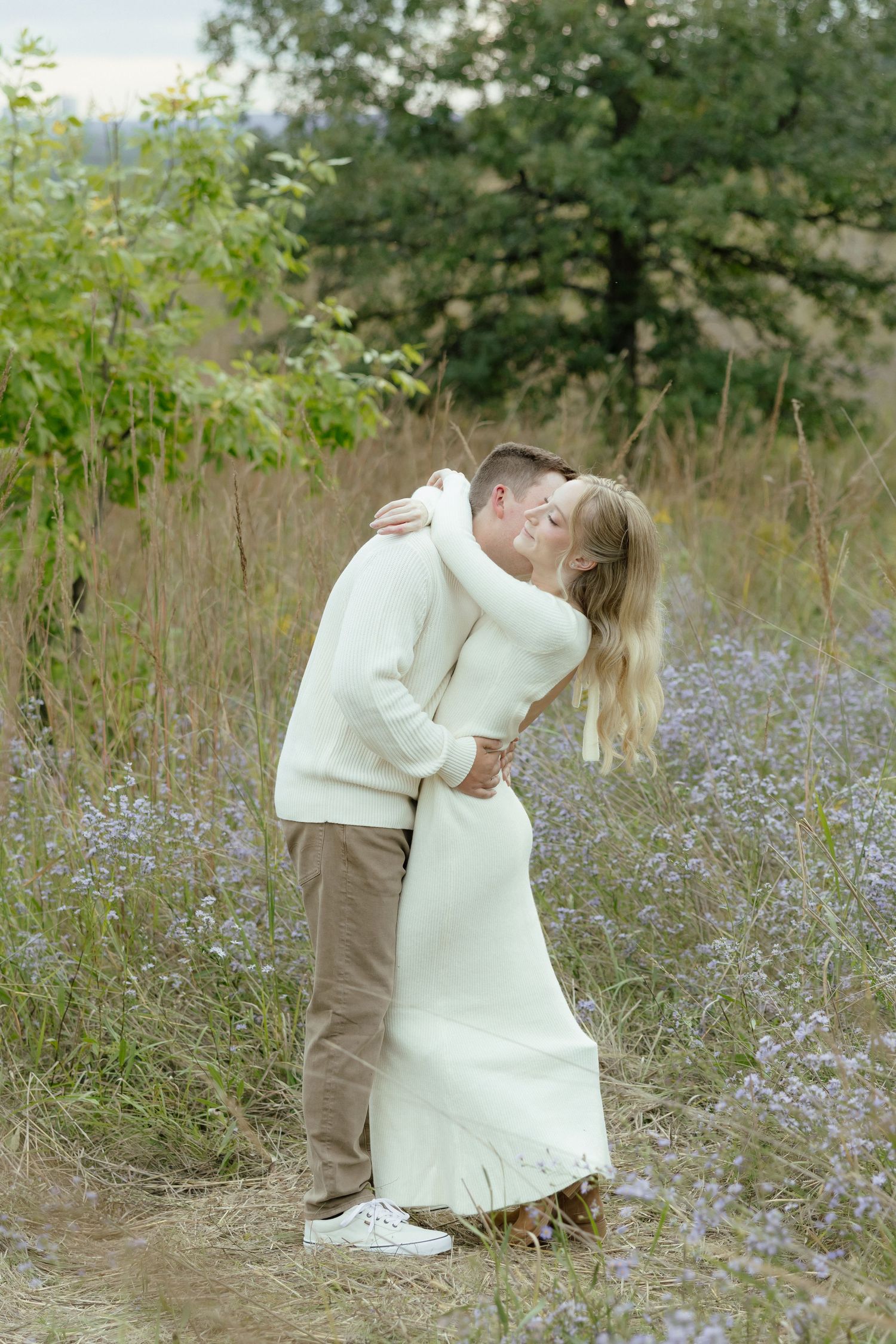 A romantic couple in white and beige embraces in a meadow filled with delicate blue wildflowers on a sunny day.