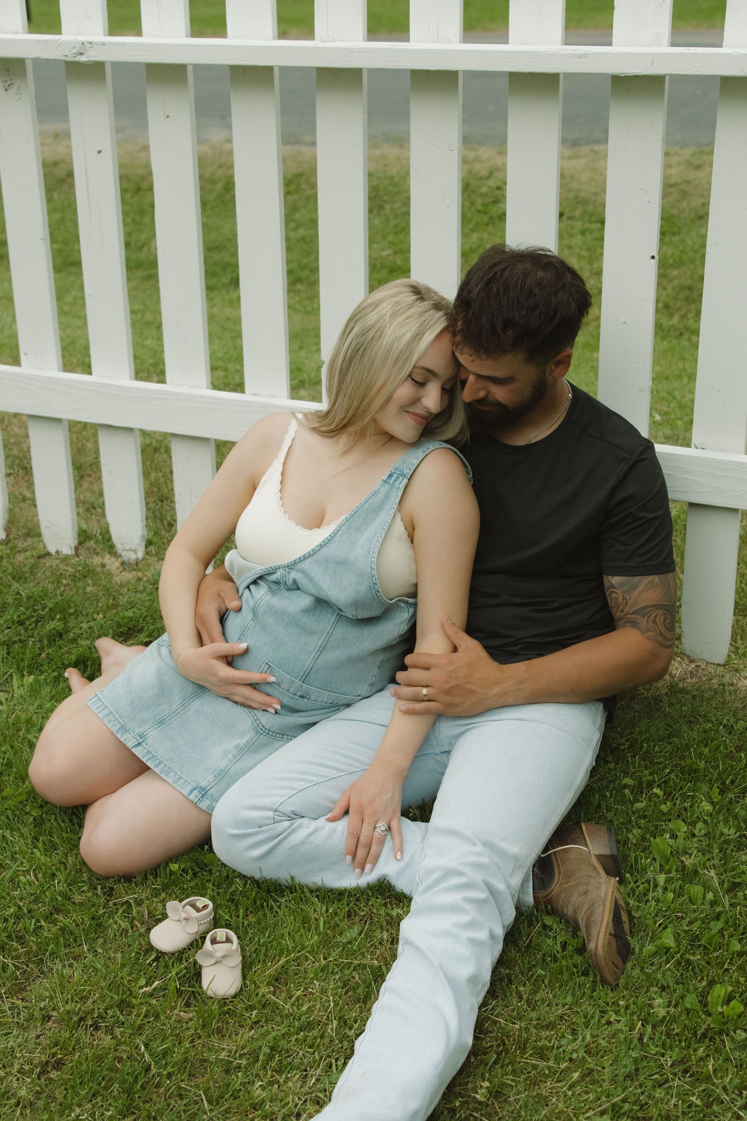 Pregnant couple sitting on grass by white picket fence, man embracing woman with baby shoes nearby.