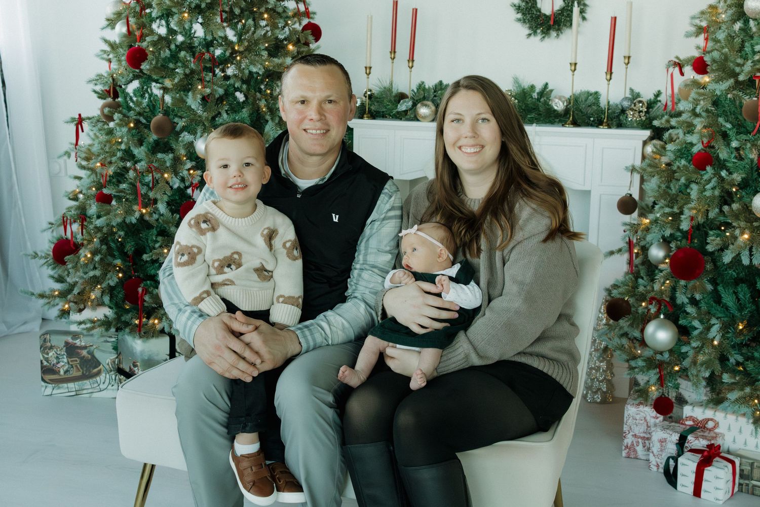 Family of four posing for Christmas portrait in front of decorated holiday trees with gifts and candles.
