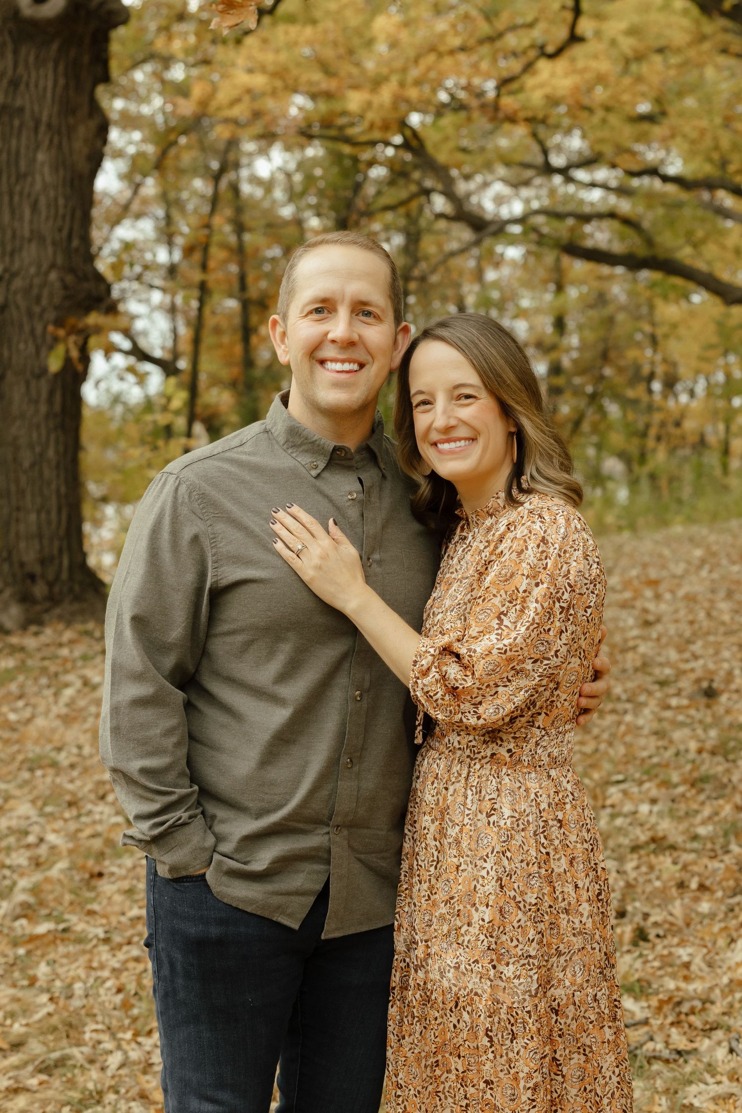 Smiling couple posing together in autumn park with colorful fall foliage, woman in floral dress, man in olive shirt.