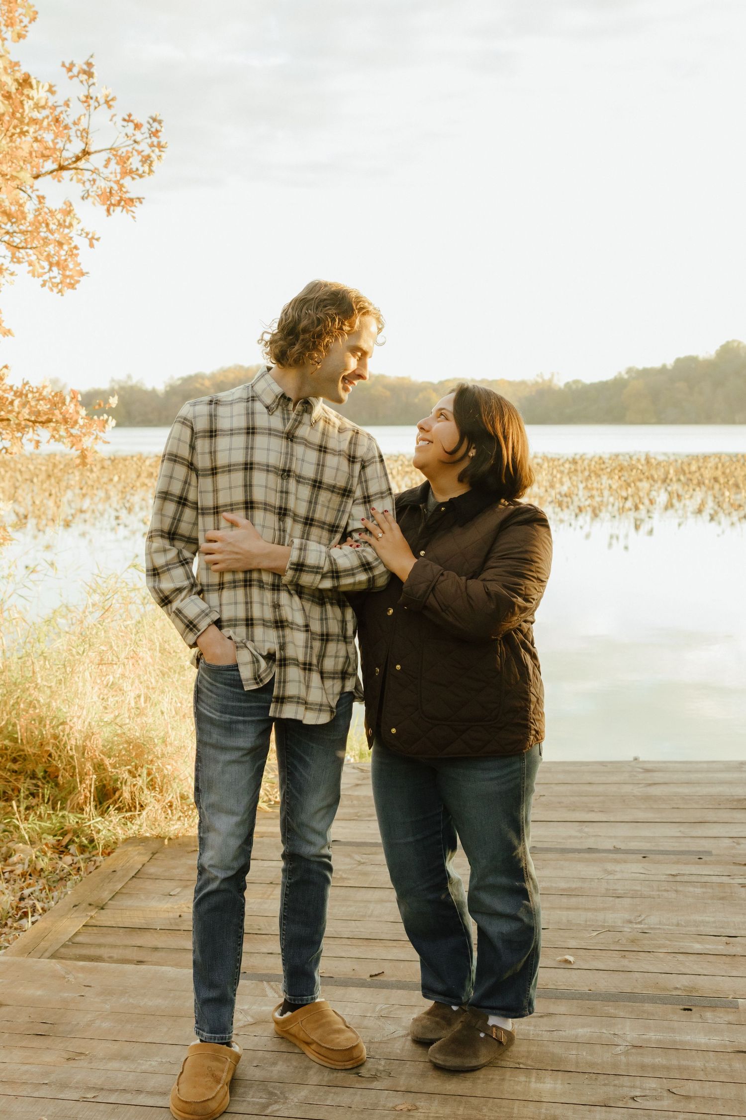 Couple sharing a warm smile on a wooden dock by a lake surrounded by golden autumn foliage.