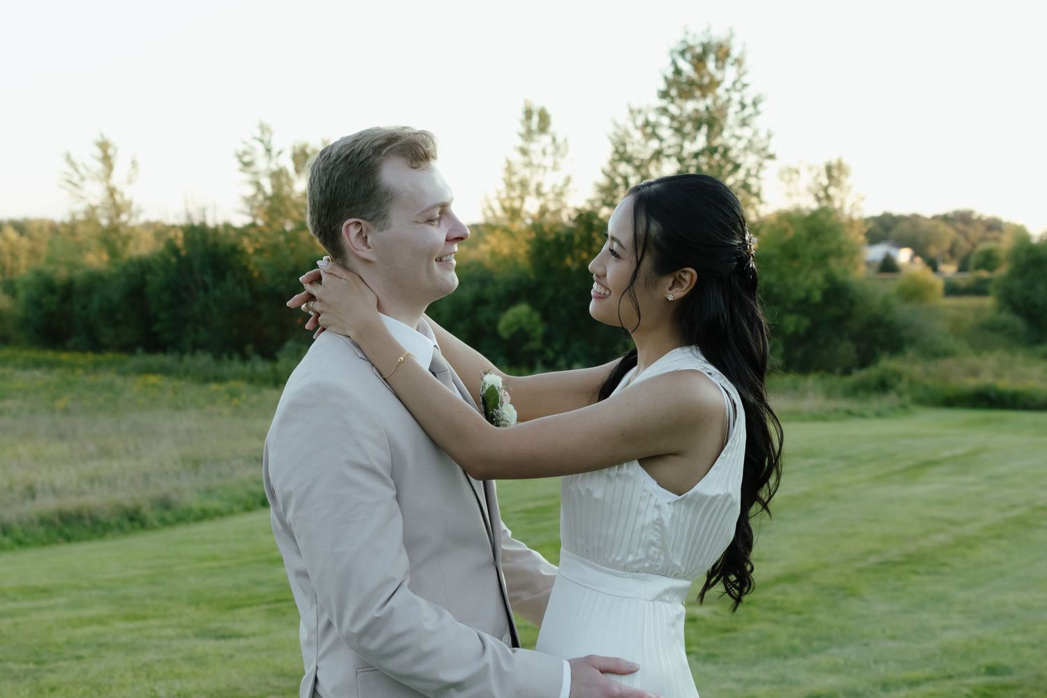 A couple in white wedding attire embraces on a grassy field during sunset.