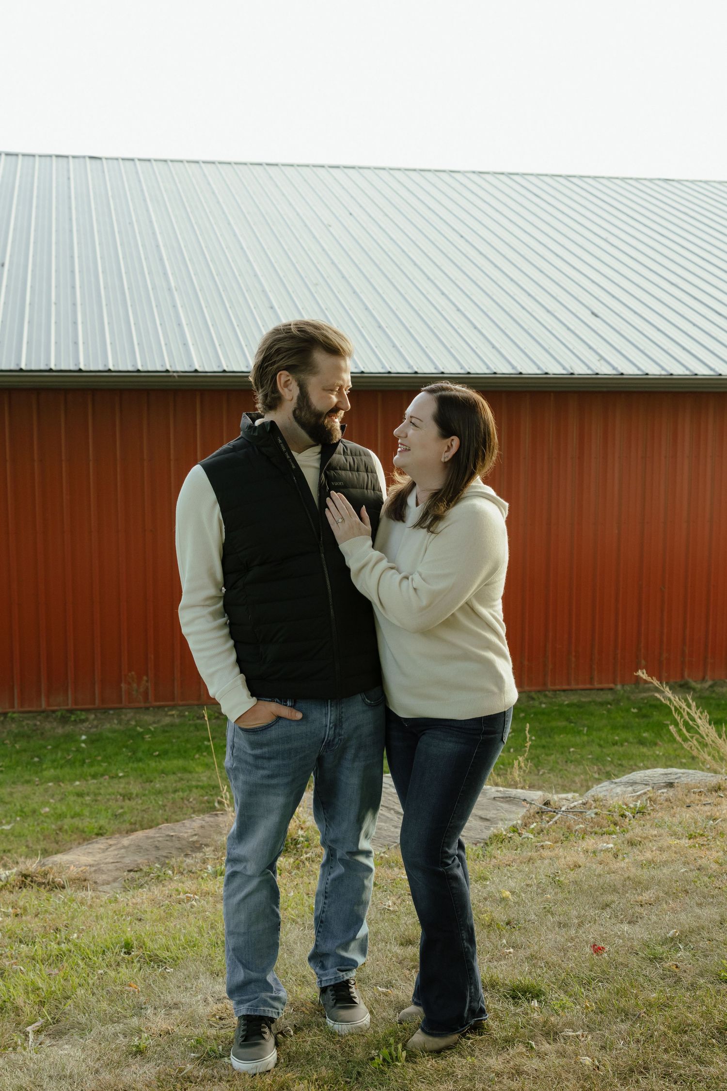A couple laughs together outside a red barn, the woman resting her hand on the man's chest in a cozy fall setting.