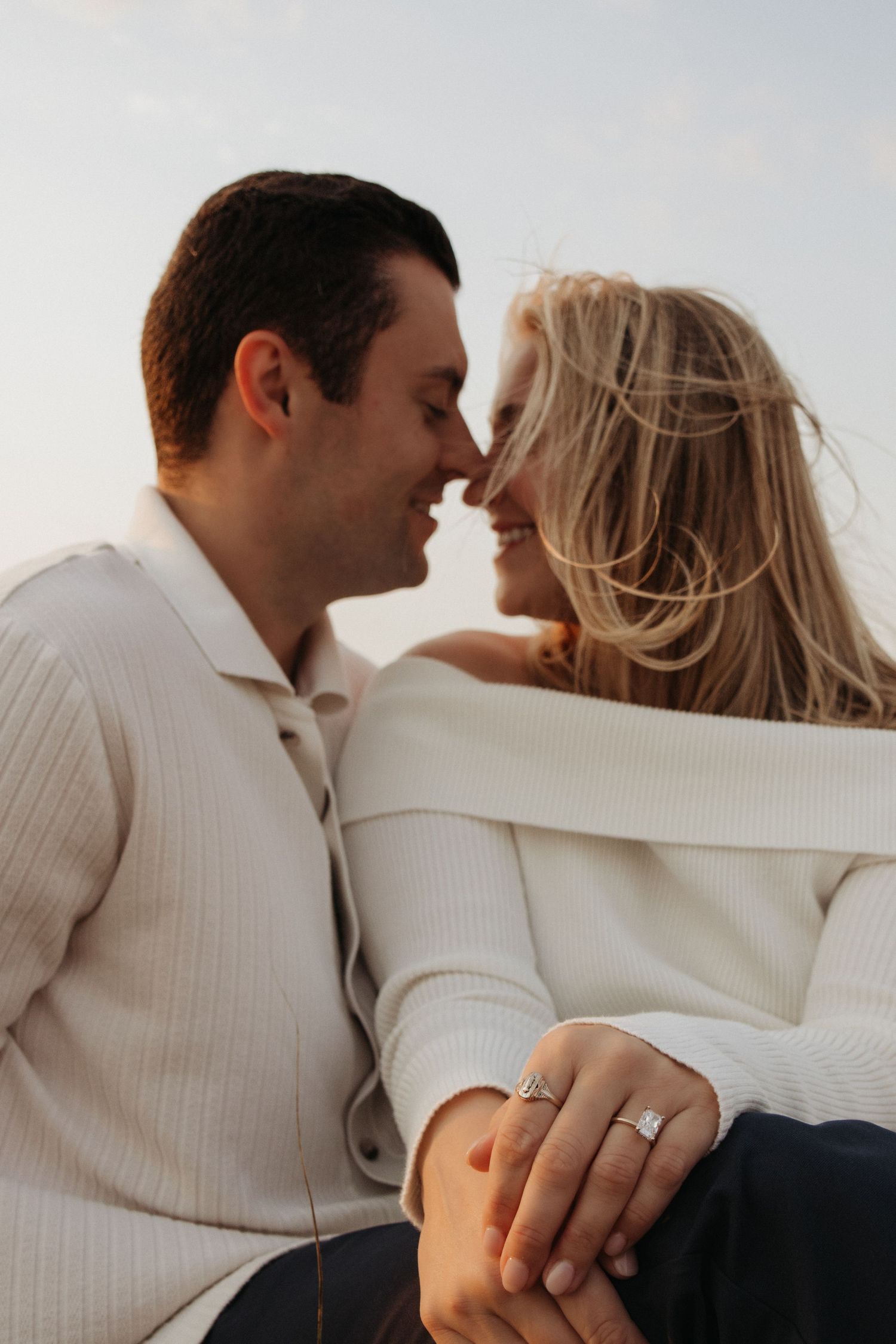 Couple in white sweaters sharing a romantic moment, woman displaying diamond engagement ring, outdoors at dusk.