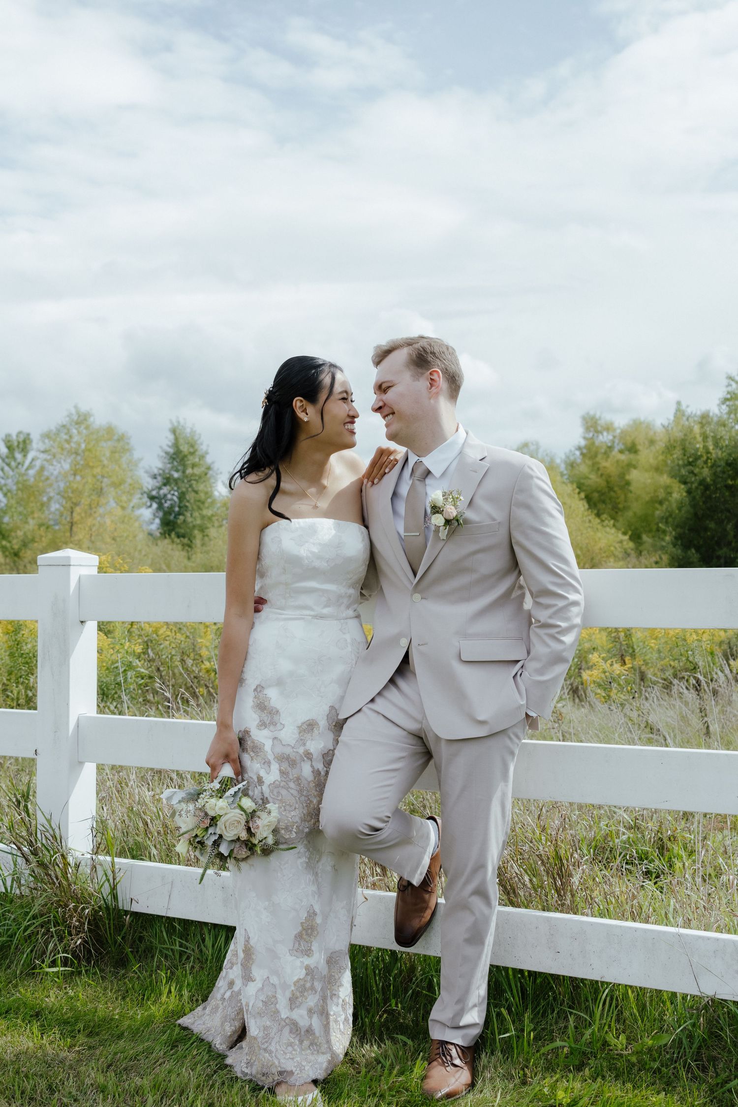 Romantic wedding couple shares intimate moments along a white fence in a rustic outdoor setting with tall grass.
