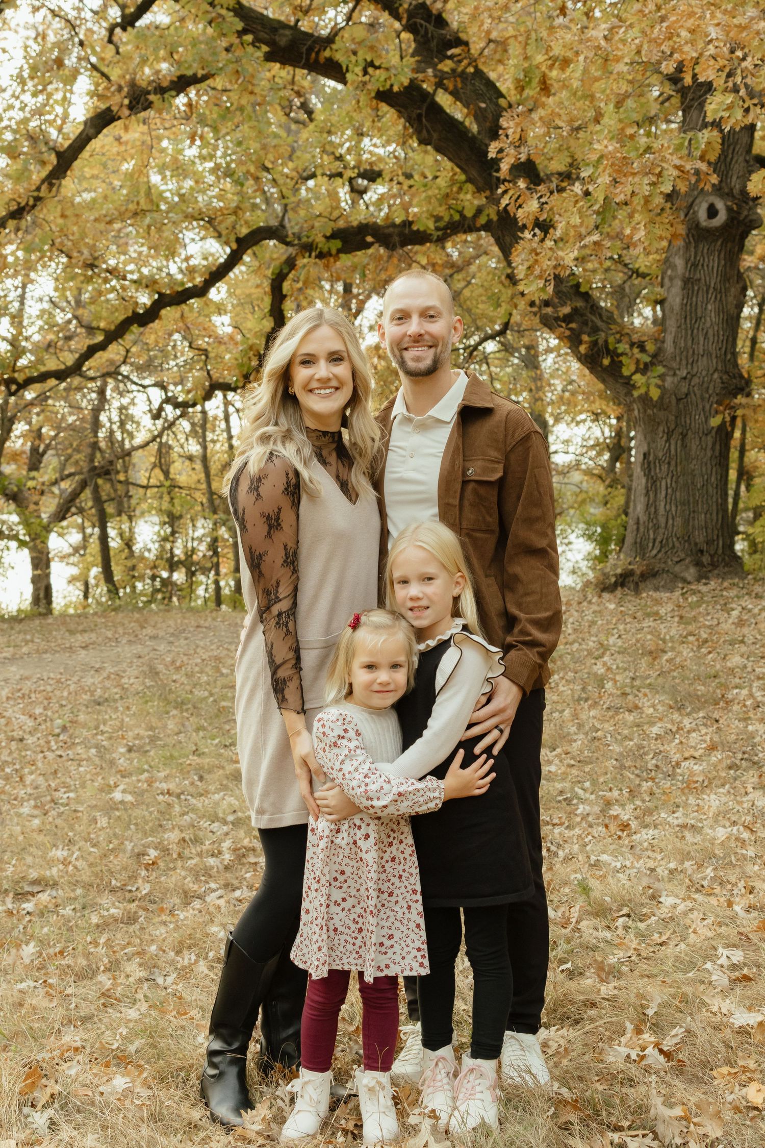 Family of four posing outdoors among autumn trees with golden foliage, parents smiling with two young daughters.
