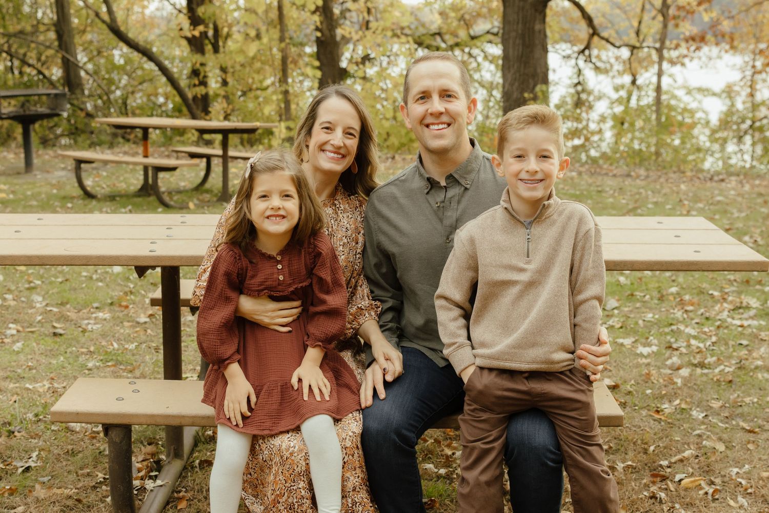 Family of four smiling together on a park bench surrounded by autumn foliage.