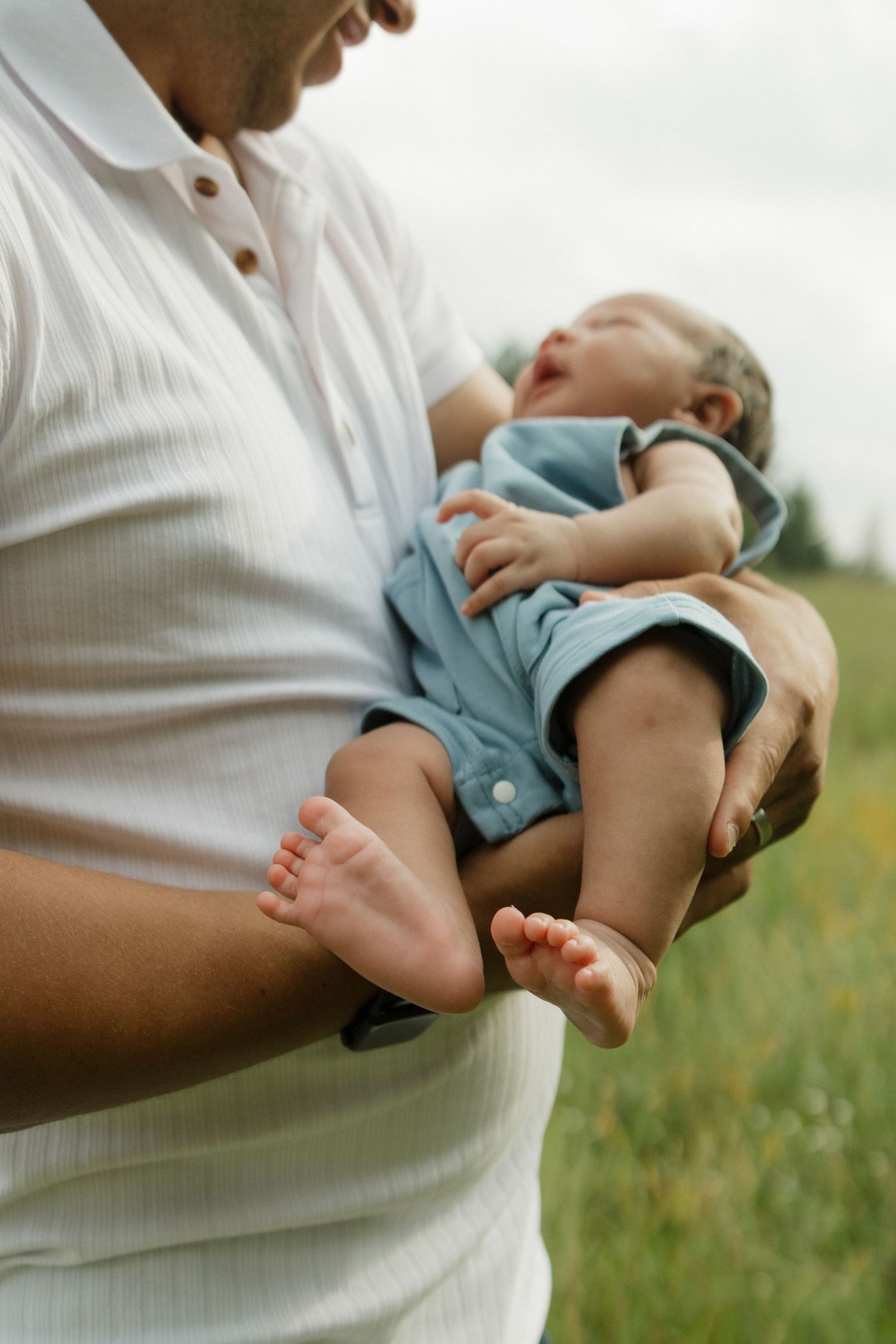 Father in white shirt cradling newborn baby in blue onesie outdoors in green field.