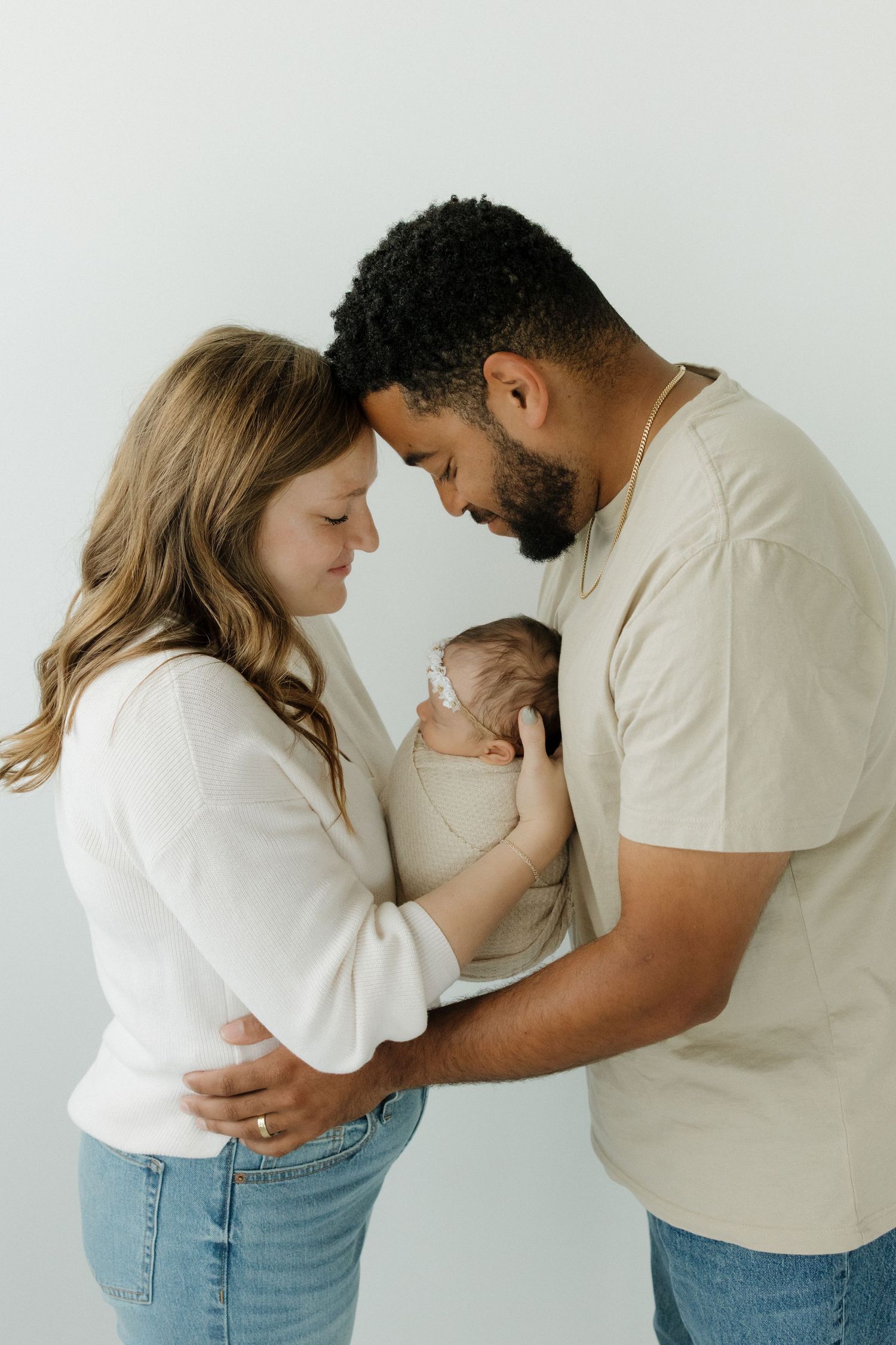 Loving parents lean foreheads together while cradling their newborn baby in a bright studio portrait.