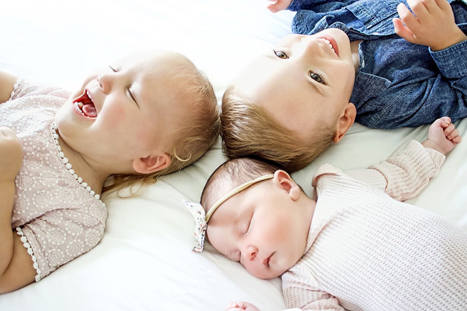 Three babies lying together on a white surface, two toddlers and a sleeping newborn in a pink headband.