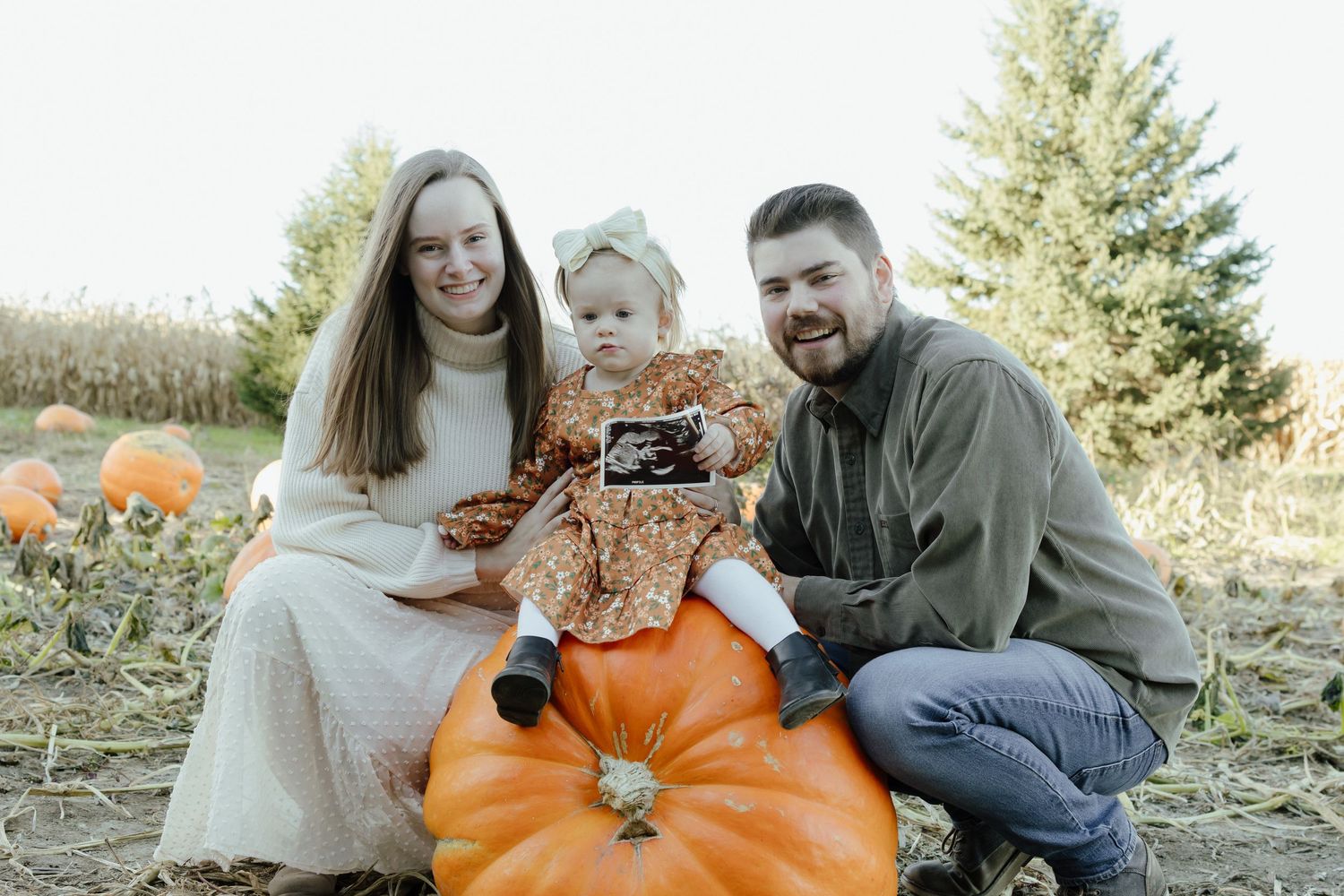 Family of three poses with ultrasound photo at a pumpkin patch, announcing a pregnancy.