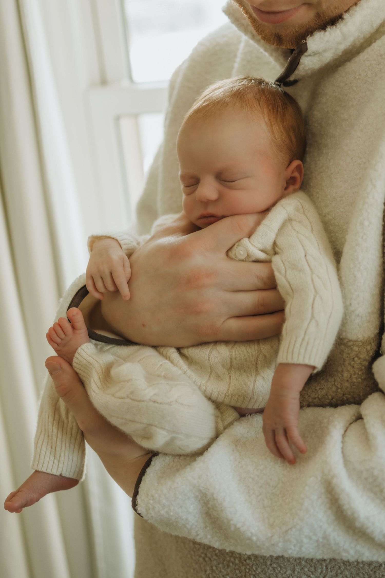 Tender moment of parent cradling sleeping newborn in white knit sweater by window.