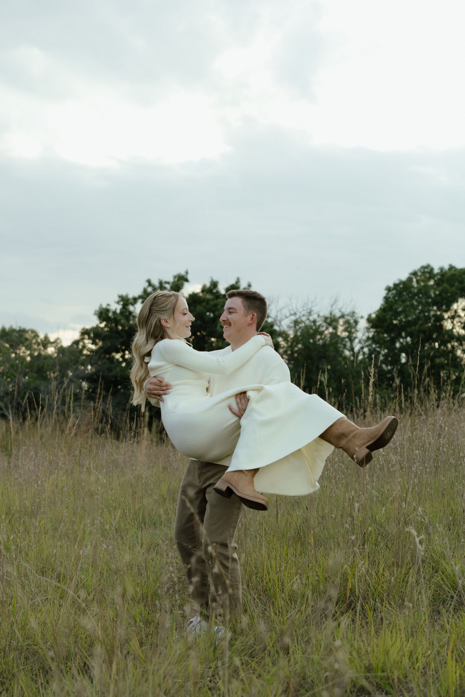 Man lifting woman in white dress in a grassy field at dusk, sharing a joyful moment outdoors.