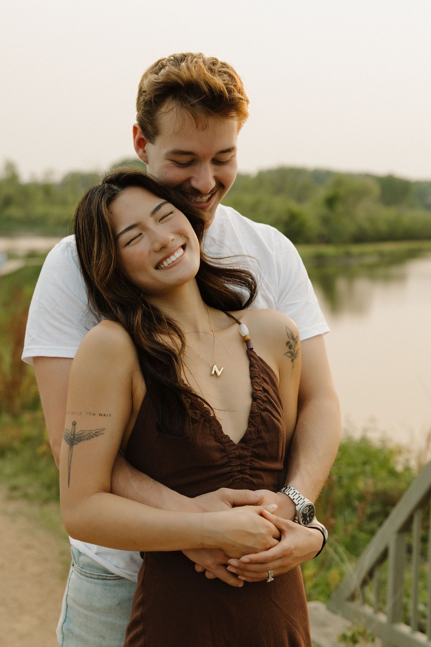 A happy couple embraces outdoors near a river, the woman smiling with eyes closed in a brown dress, man in white tee.