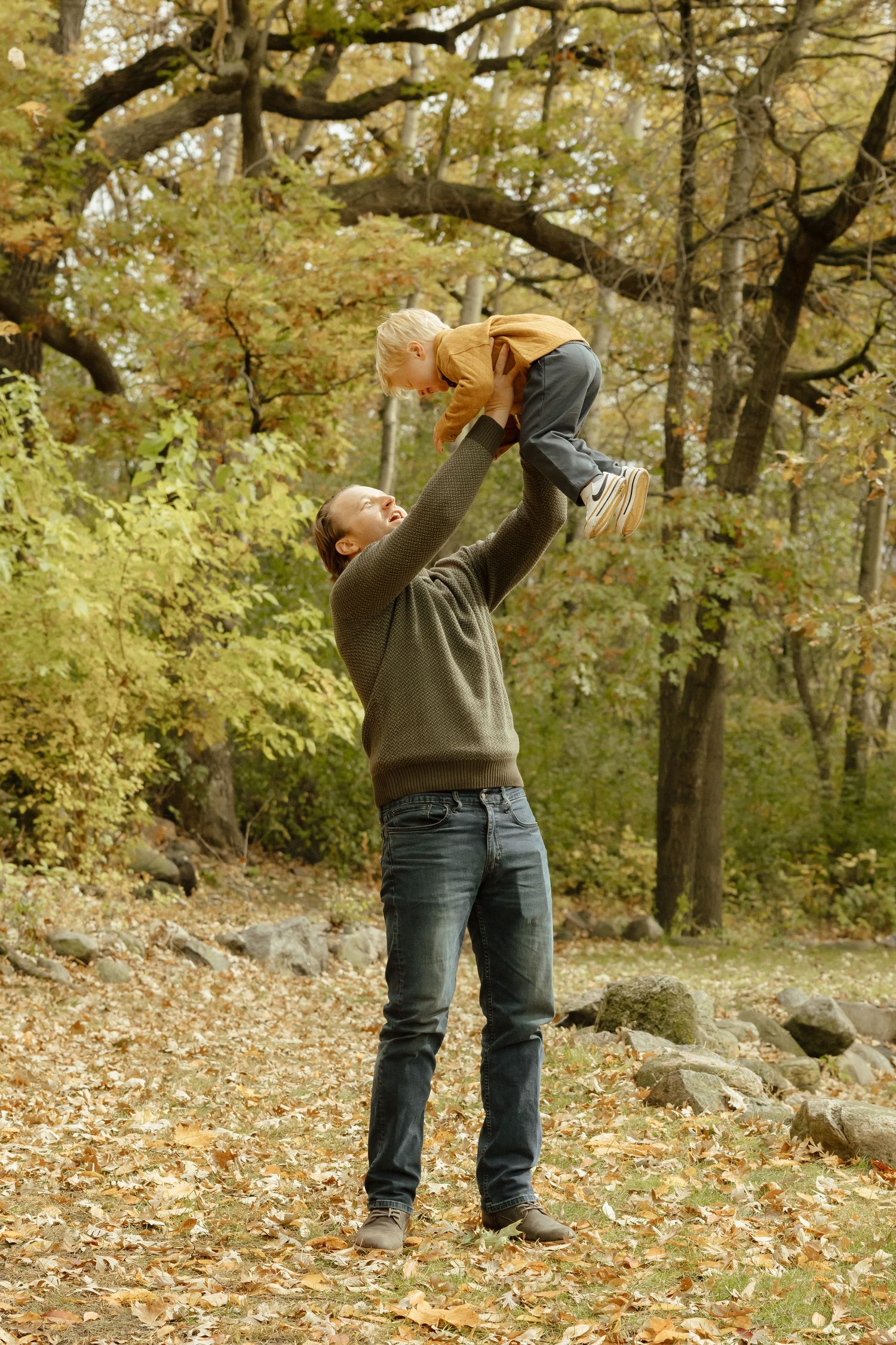 Father joyfully lifting toddler high in autumn forest park, surrounded by golden fall foliage and fallen leaves.