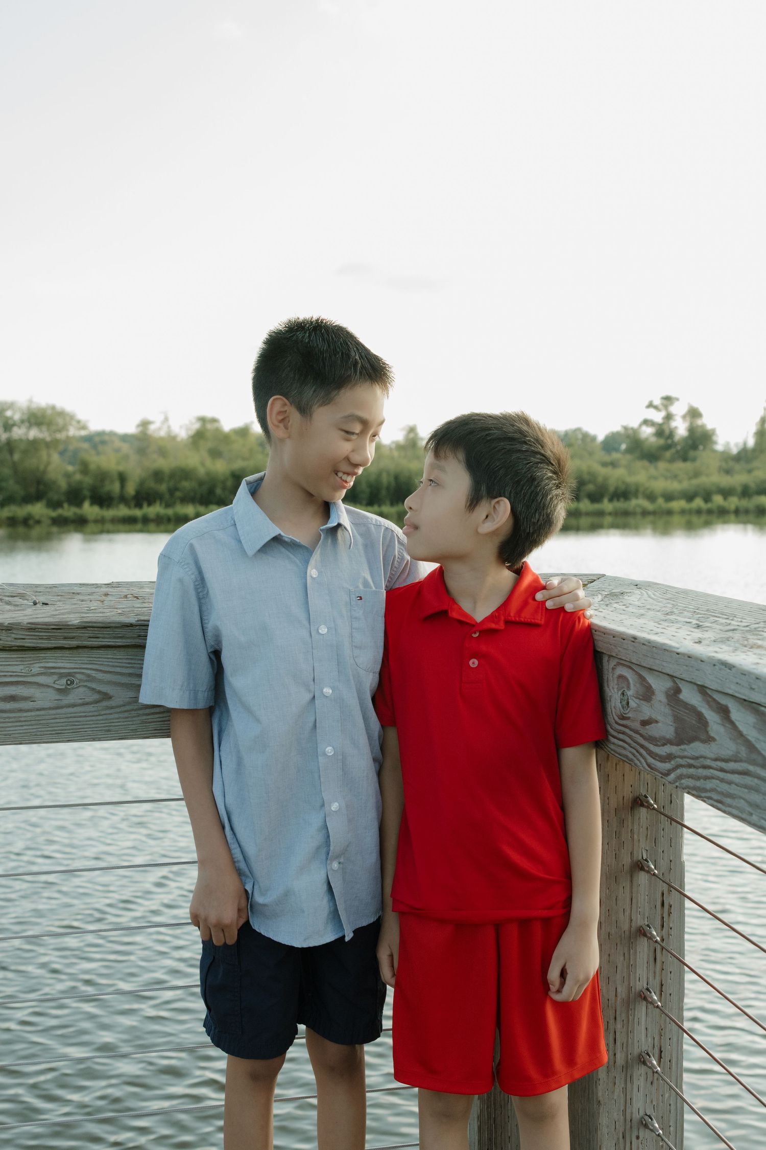Two young boys smiling and standing together on a wooden dock near a lake, one in blue shirt and one in red outfit.