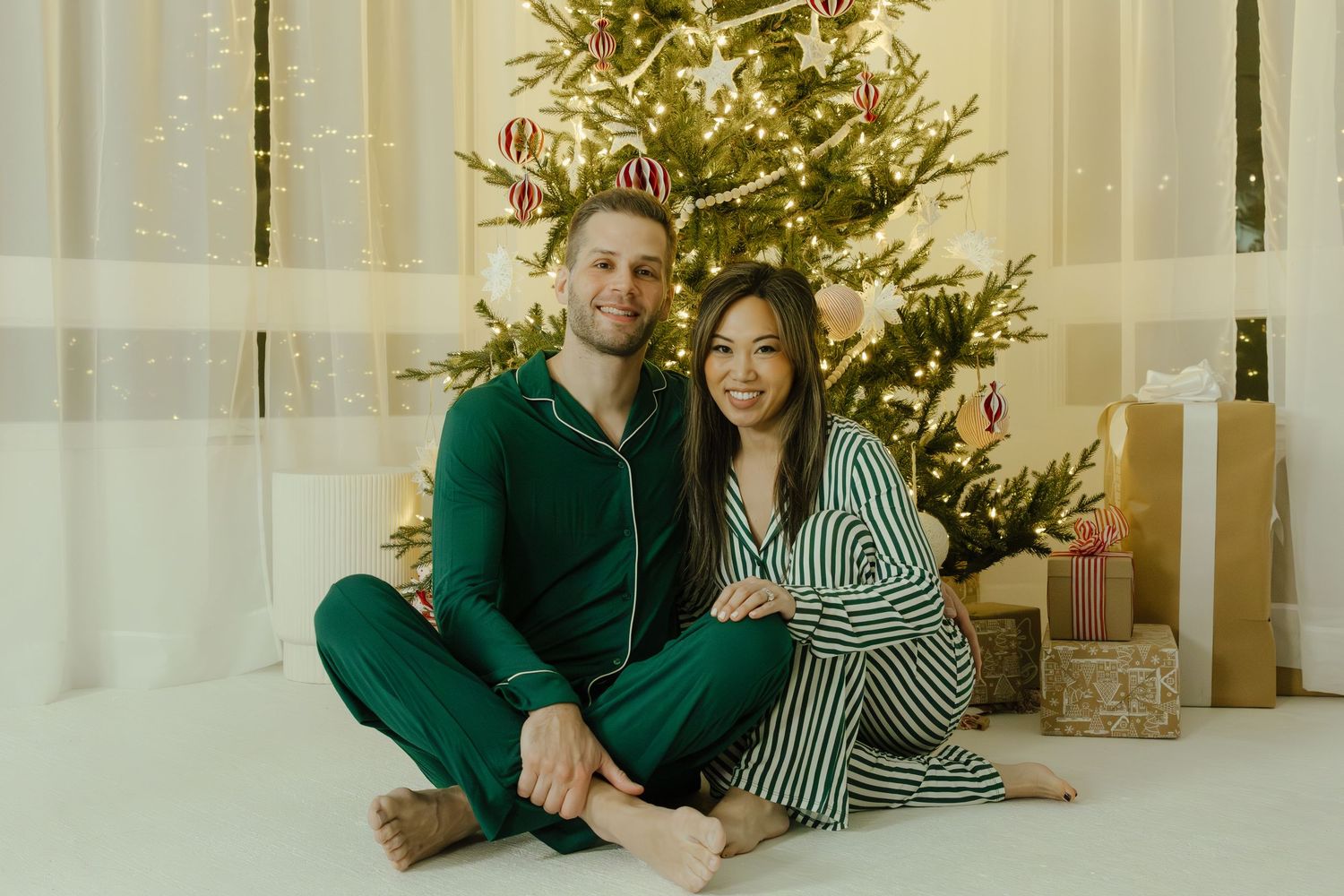 Couple in matching green pajamas sitting together in front of decorated Christmas tree with gifts.