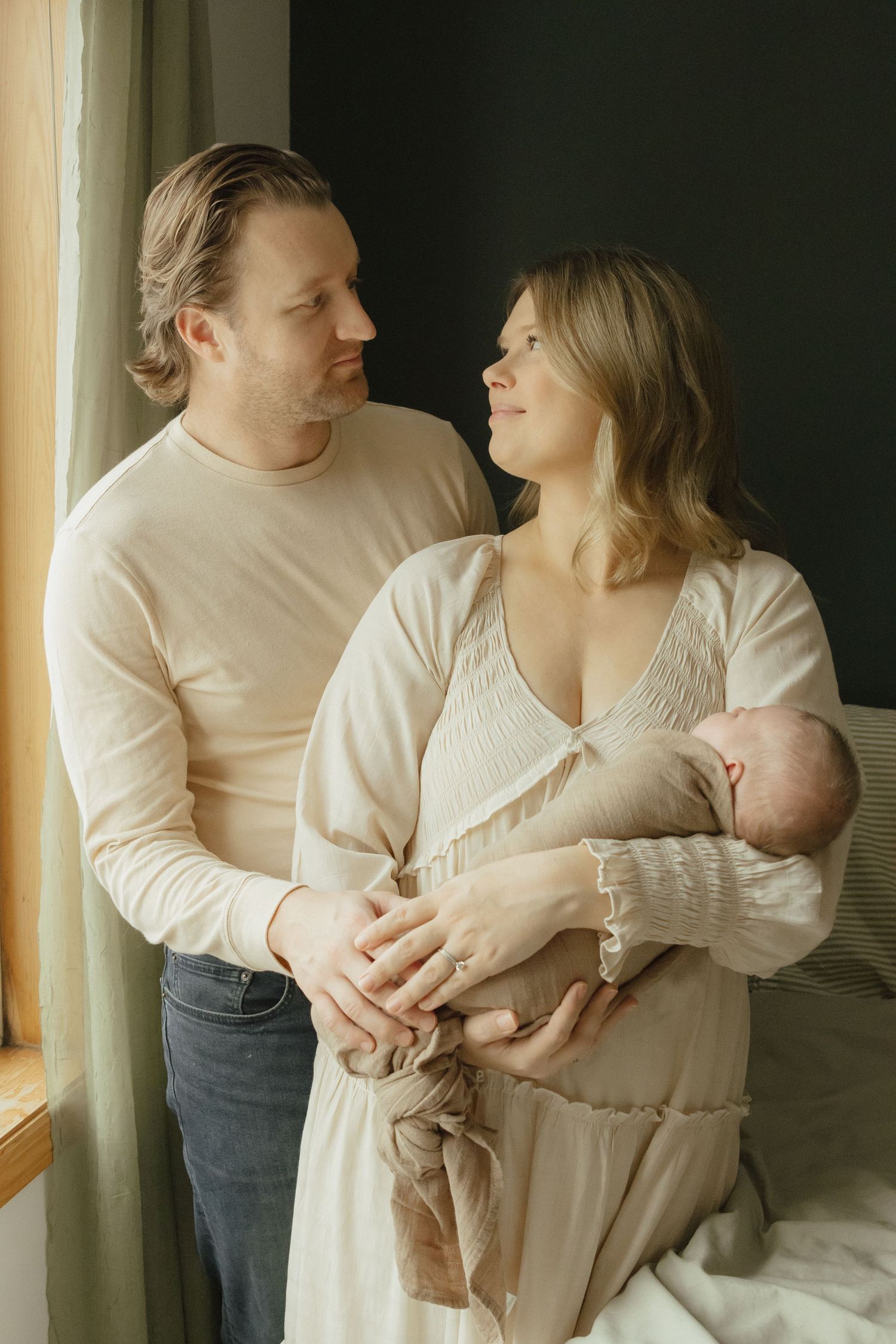 A couple in neutral tones gazes at each other while tenderly holding their newborn baby near a sunlit window.