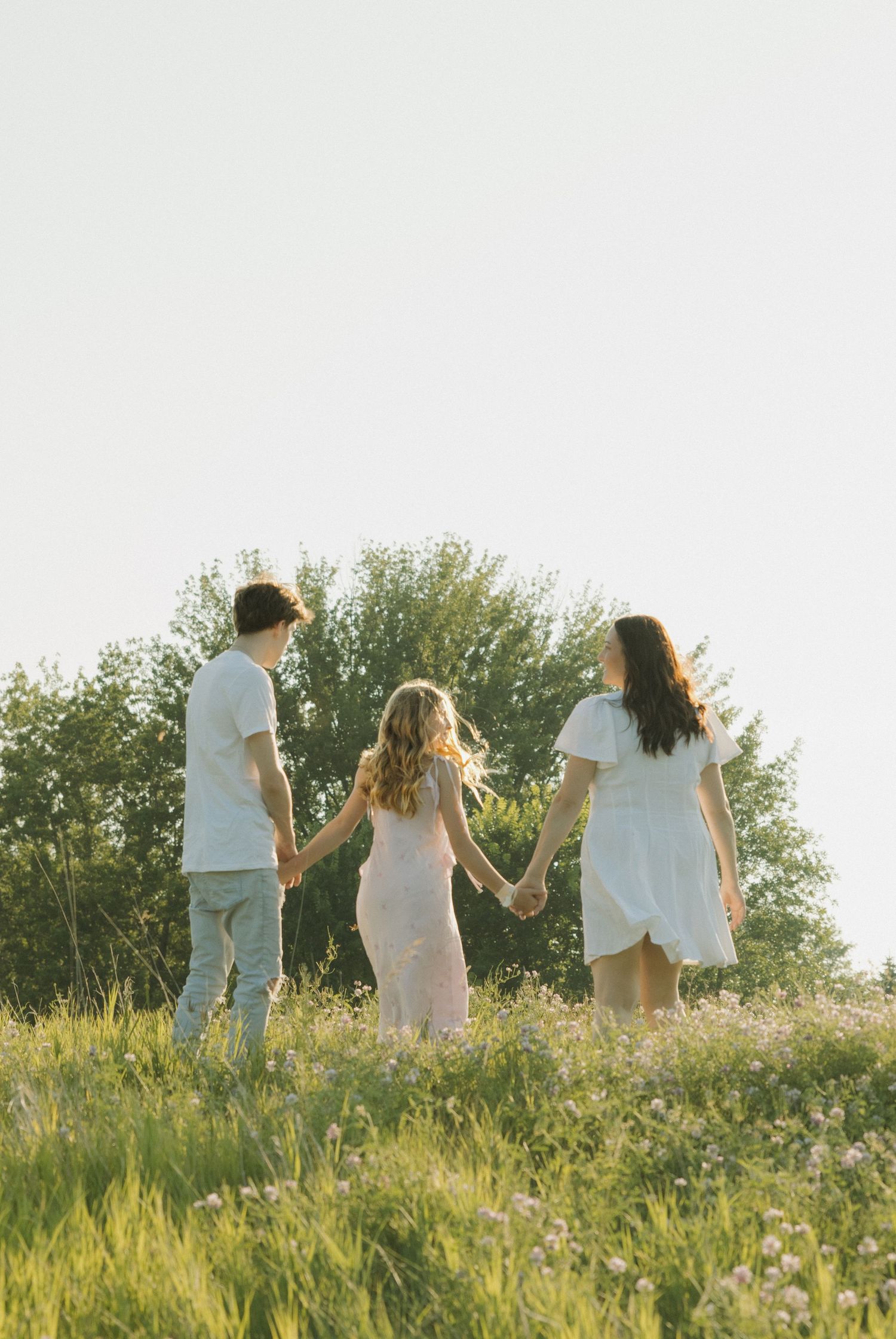 Three people holding hands walking through a sunny green meadow with wildflowers and trees in the background.