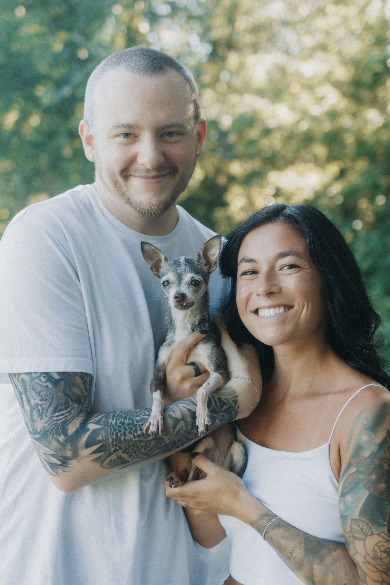A couple poses outdoors with their small gray dog, both wearing white shirts.