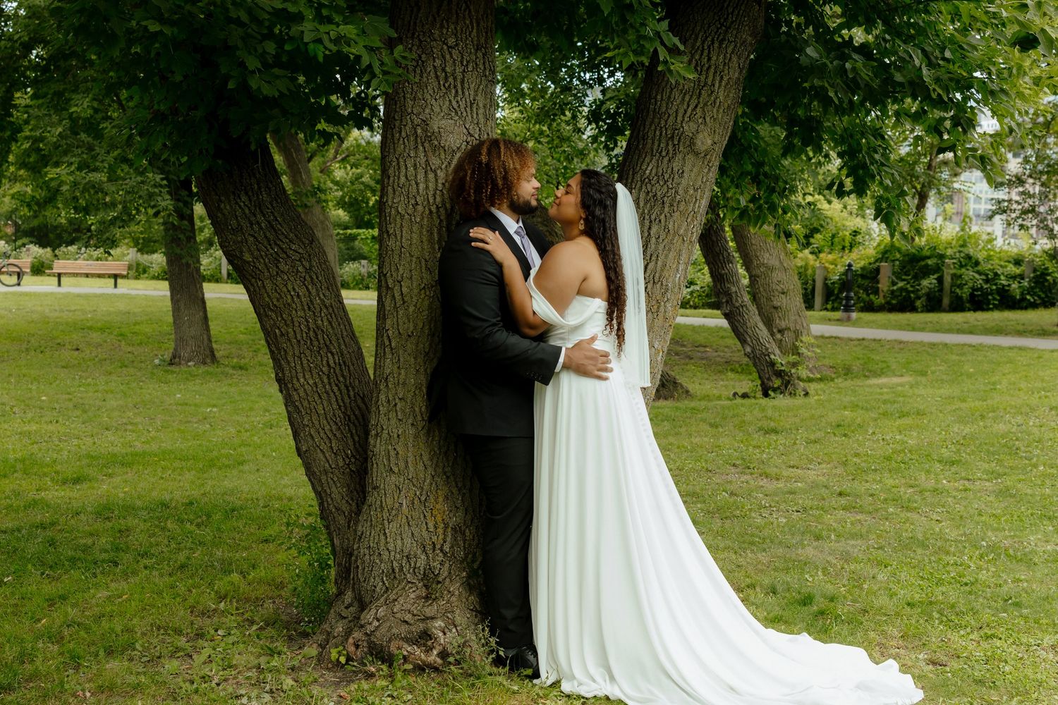 A couple in wedding attire share a romantic moment against a large tree trunk in a lush green park setting.