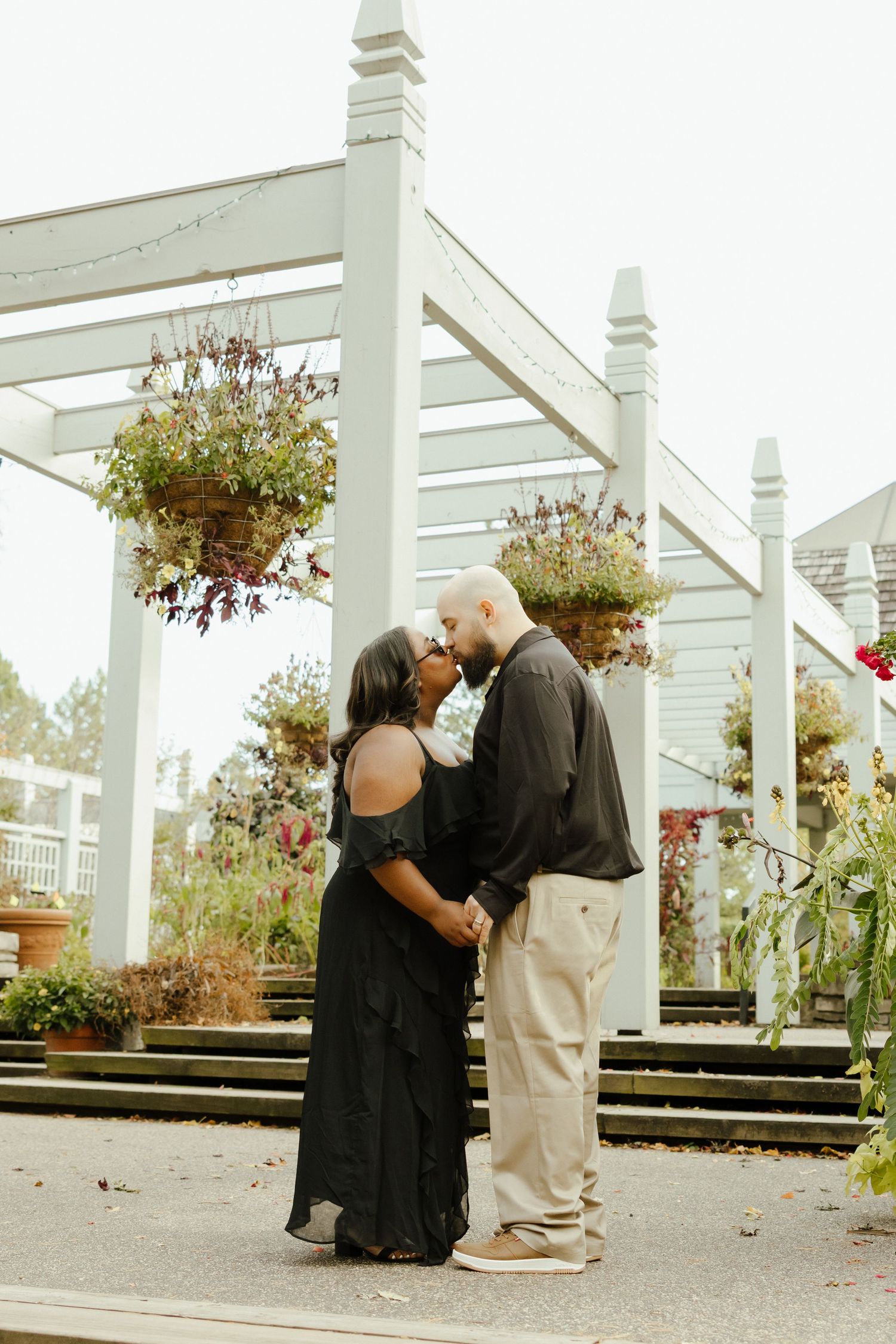 Couple sharing a romantic kiss under a white pergola with hanging flower baskets in a garden setting.