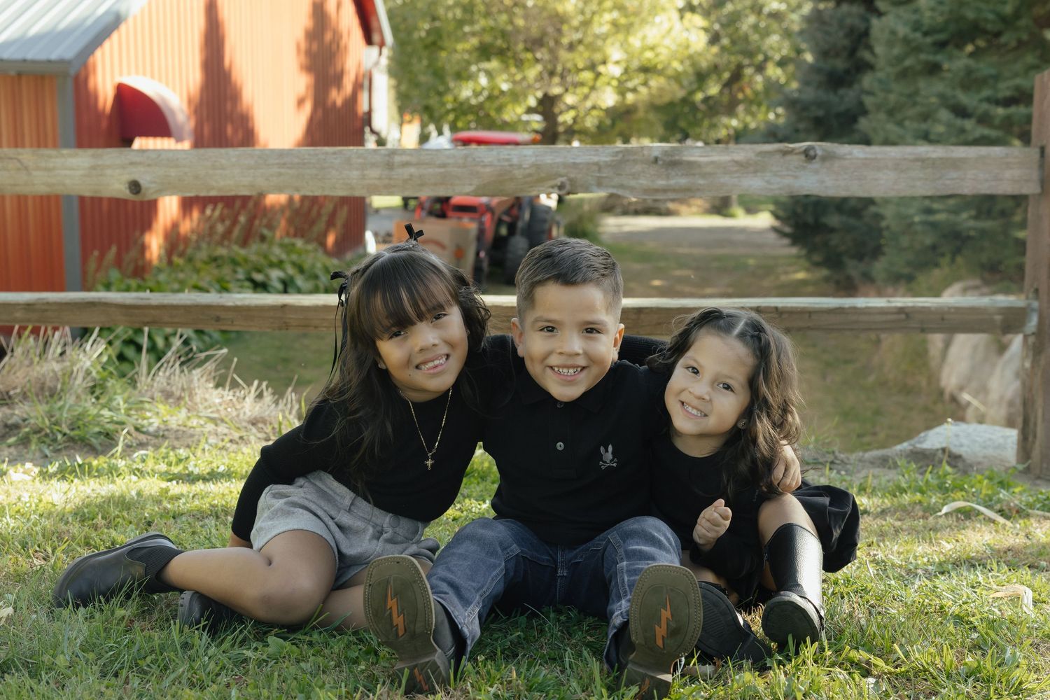 Three smiling children in black shirts sit together on grass near a wooden fence and red barn outdoors.