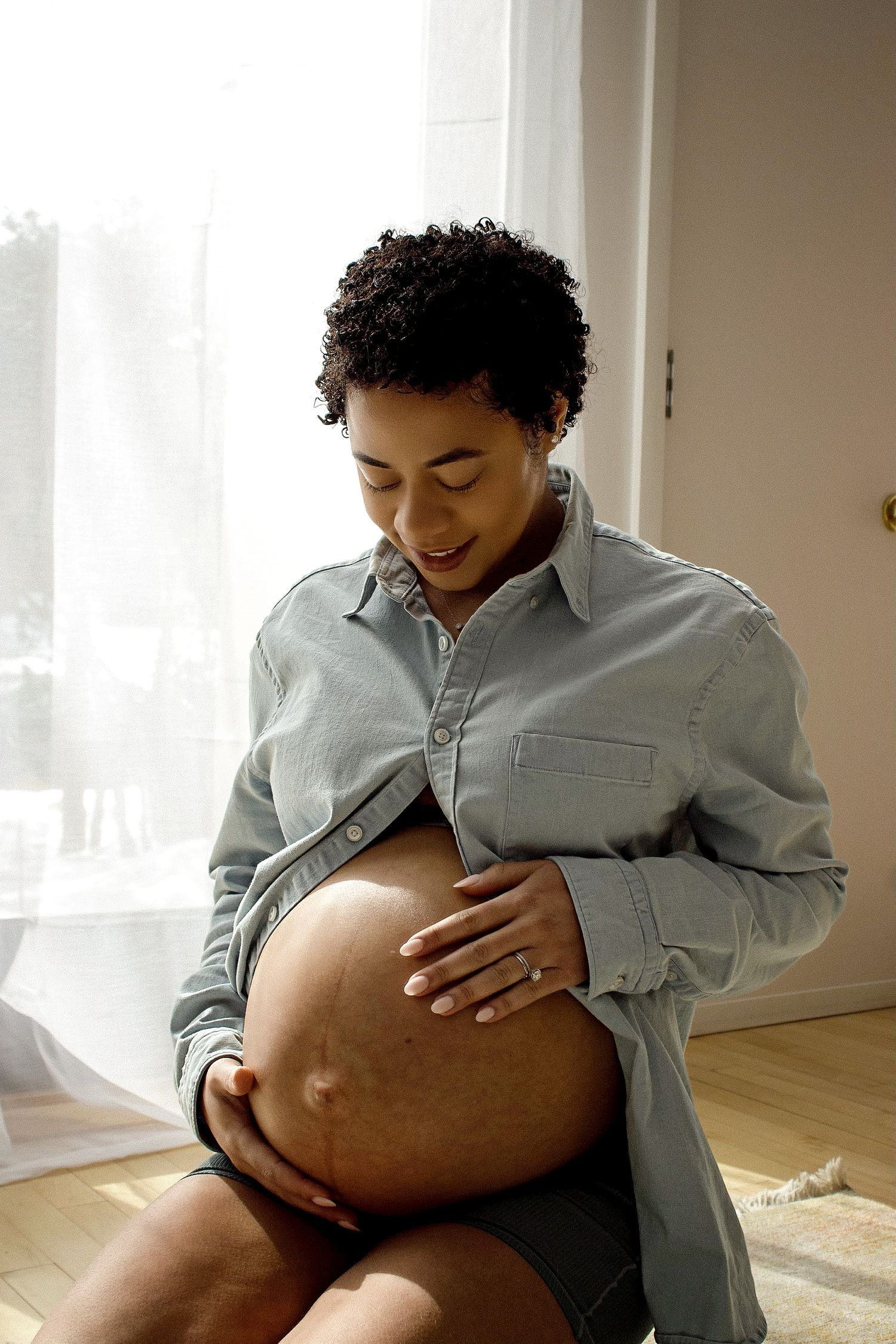 Pregnant woman in gray shirt sitting and lovingly cradling her baby bump near a bright window.