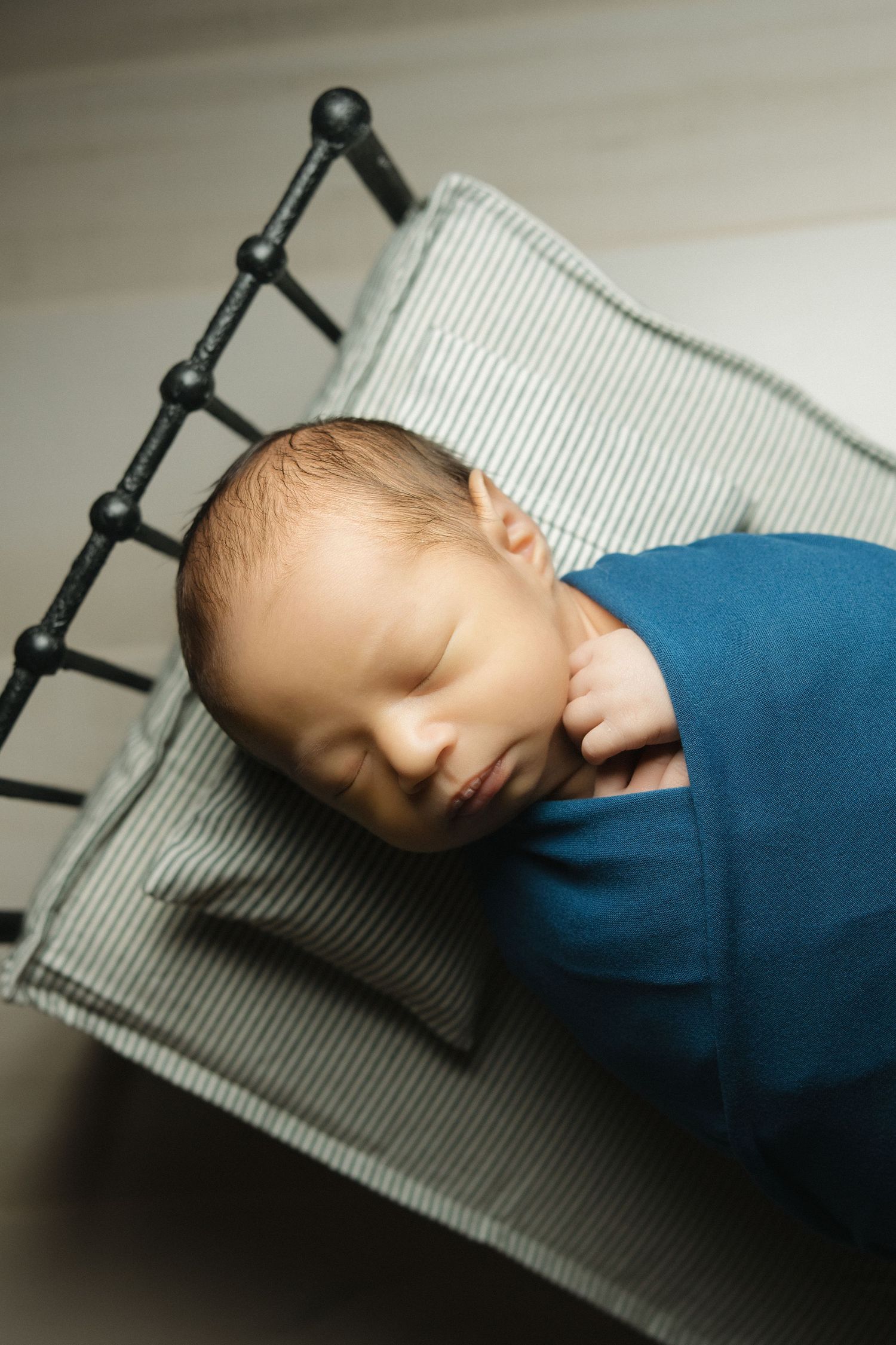 Newborn baby swaddled in blue wrap sleeping peacefully on a miniature striped bed with black metal frame.
