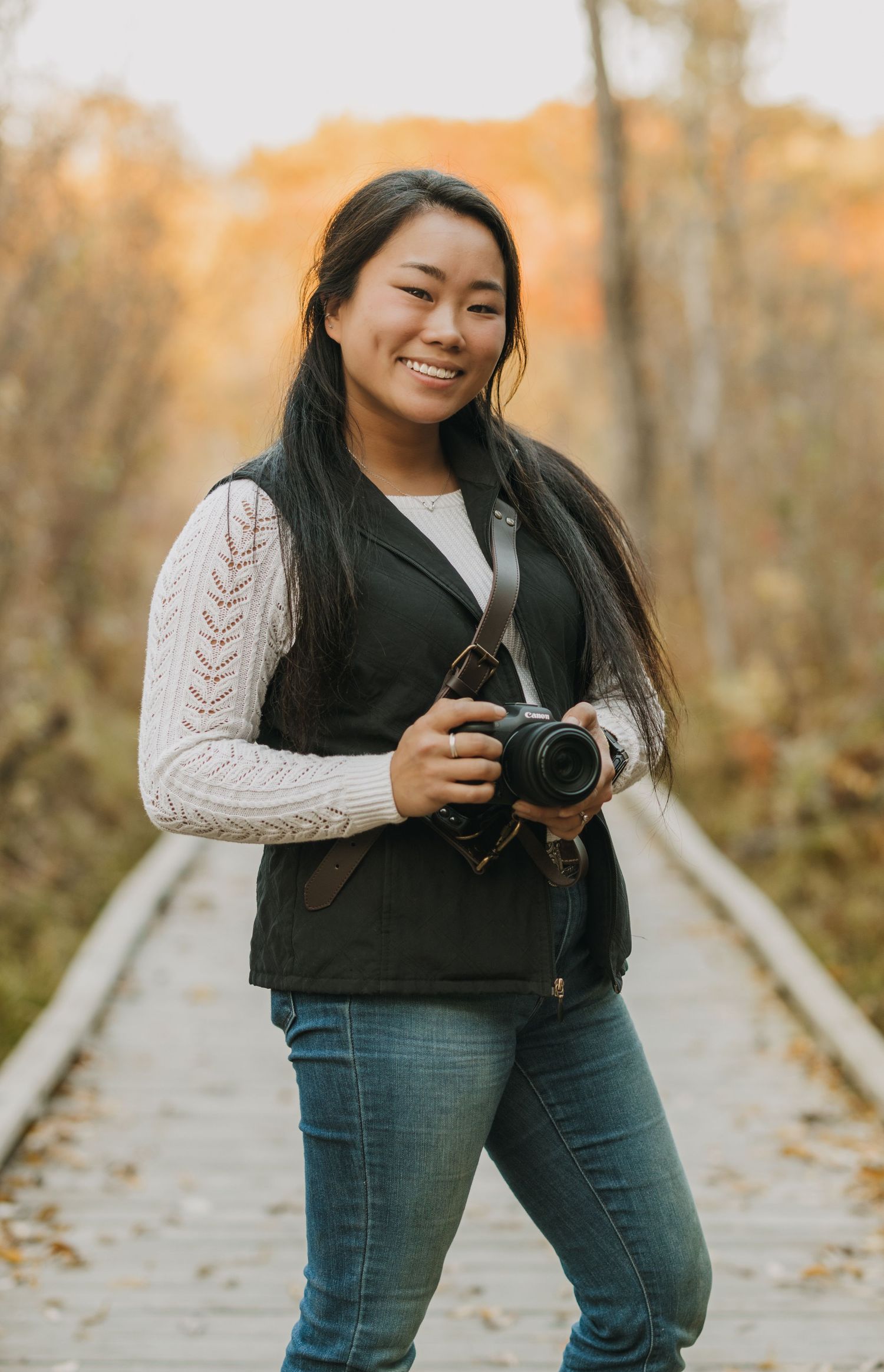 Person in knit sweater and jeans holding camera on leaf-covered path during autumn.