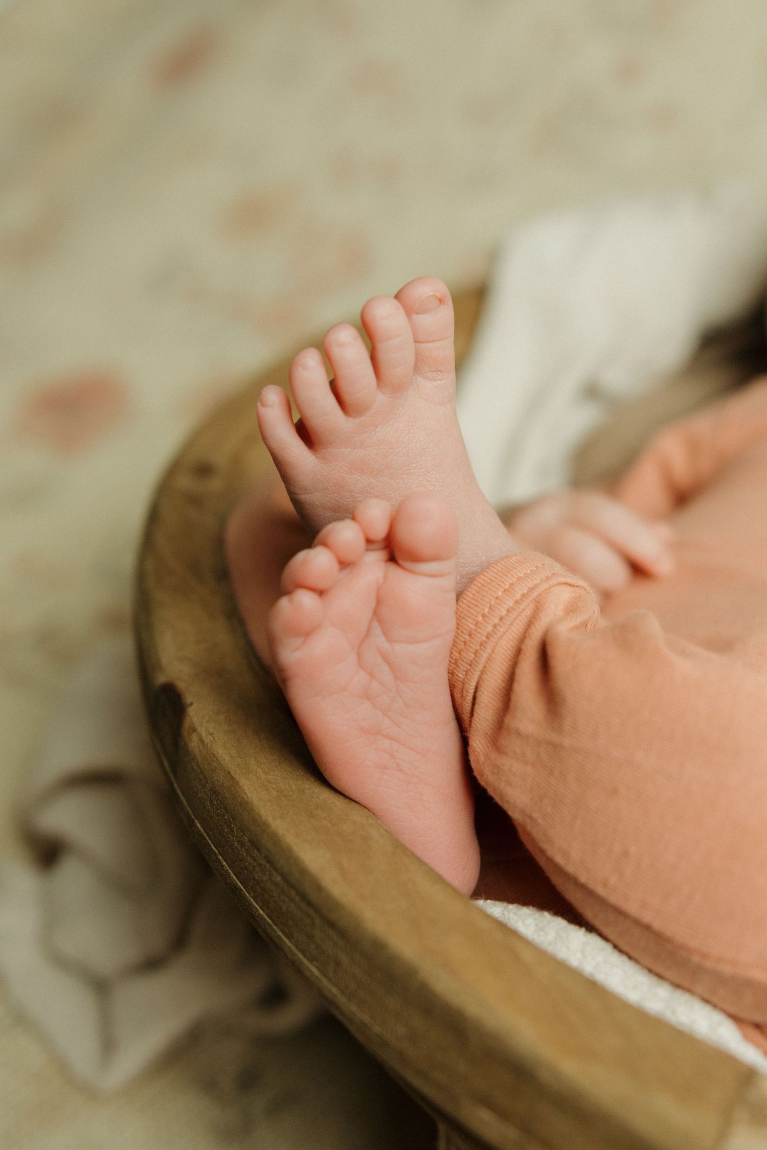 Close-up of tiny newborn baby feet in a wooden bowl, wearing a peach outfit on a soft white blanket.