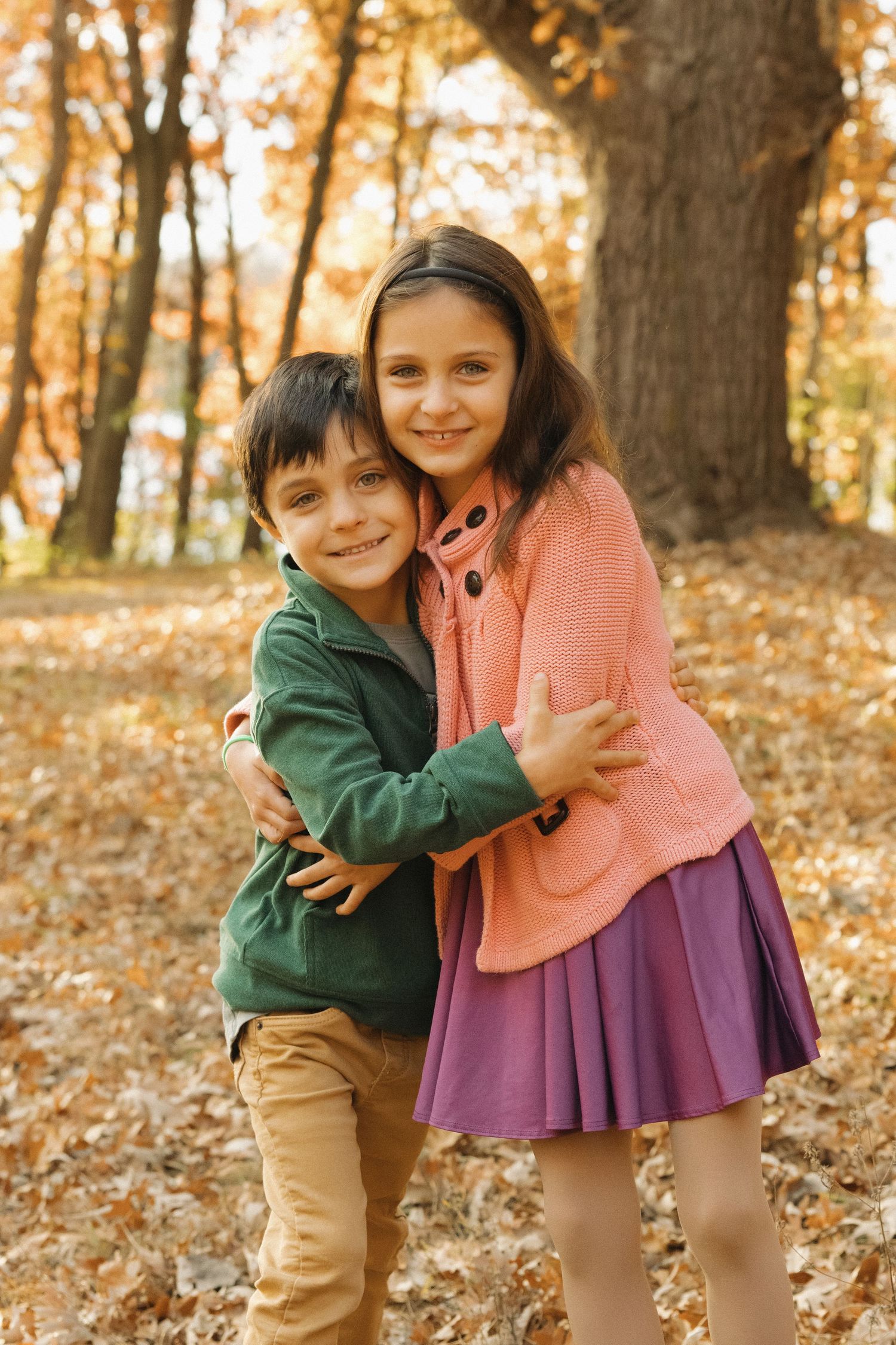 Two happy children hugging in an autumn park surrounded by golden fallen leaves and tall oak trees.