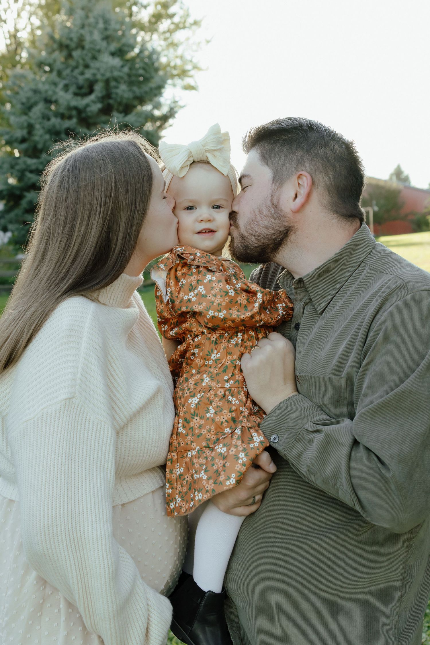 Parents kissing their smiling baby girl in a floral dress with a bow outdoors near green trees.