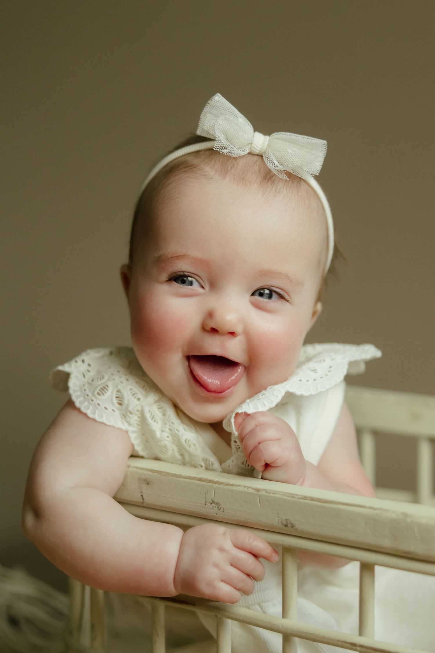 A happy baby wearing a white dress and bow peeks over a white crib rail with a delightful expression.