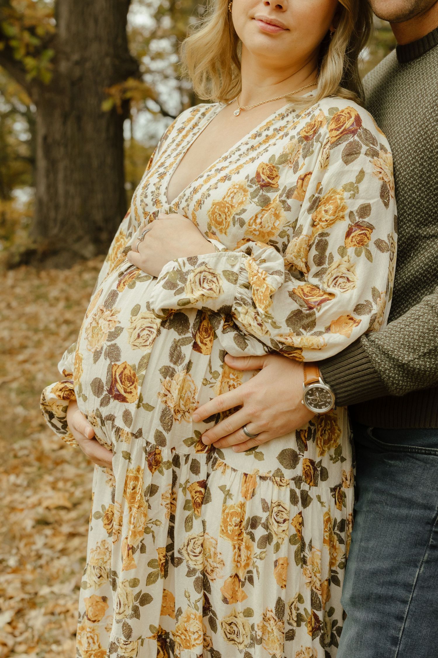 Pregnant woman in floral dress embraced by partner during autumn maternity photoshoot in wooded setting.