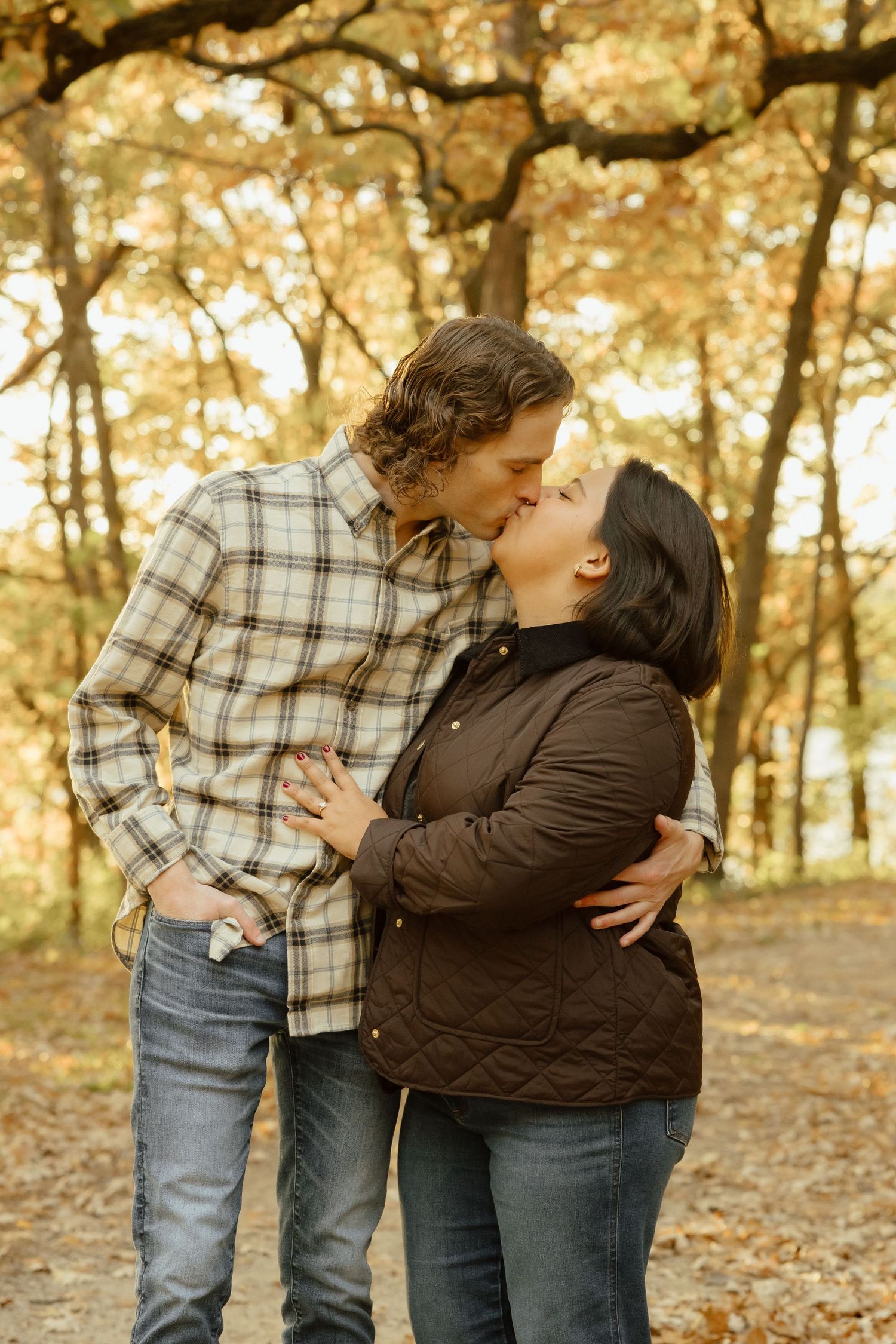 Couple sharing a romantic kiss in autumn forest, wearing casual flannel and quilted jackets among golden fall foliage.