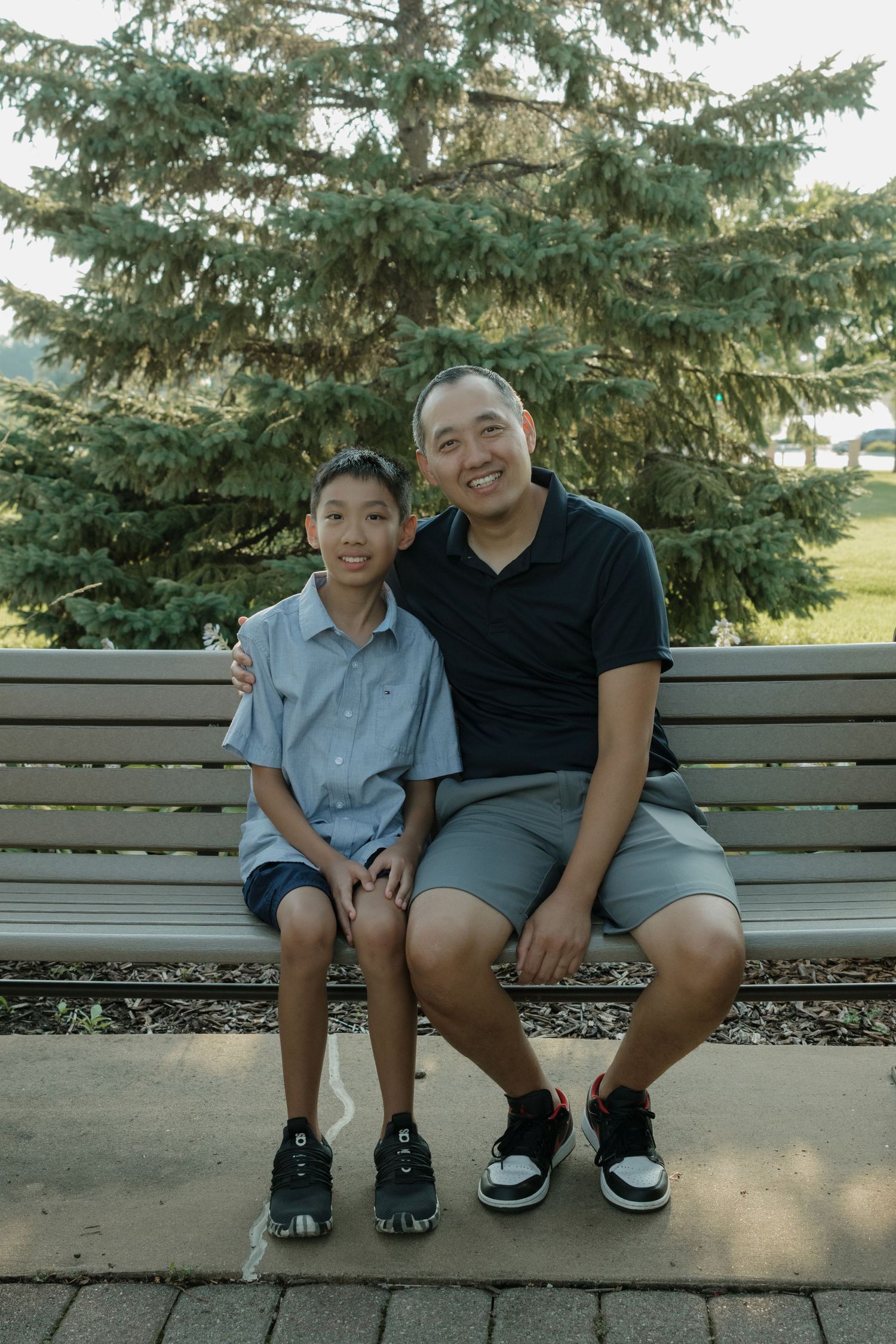 A man and young boy smile together on a park bench with evergreen trees in the background.