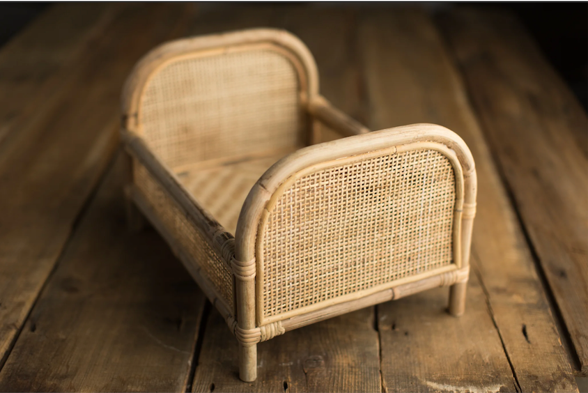 Miniature rattan cane bed with wooden frame, handcrafted newborn photography prop on rustic wood surface.