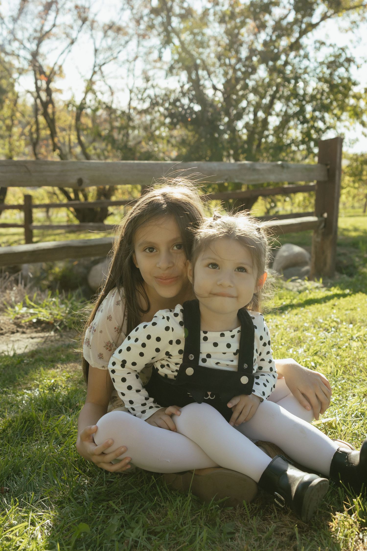 Two young sisters in polka dot tops sit together on grass near a wooden fence outdoors.