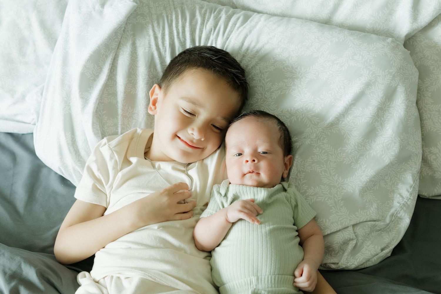 Smiling young boy lying next to sleeping newborn baby sibling on white bedding.