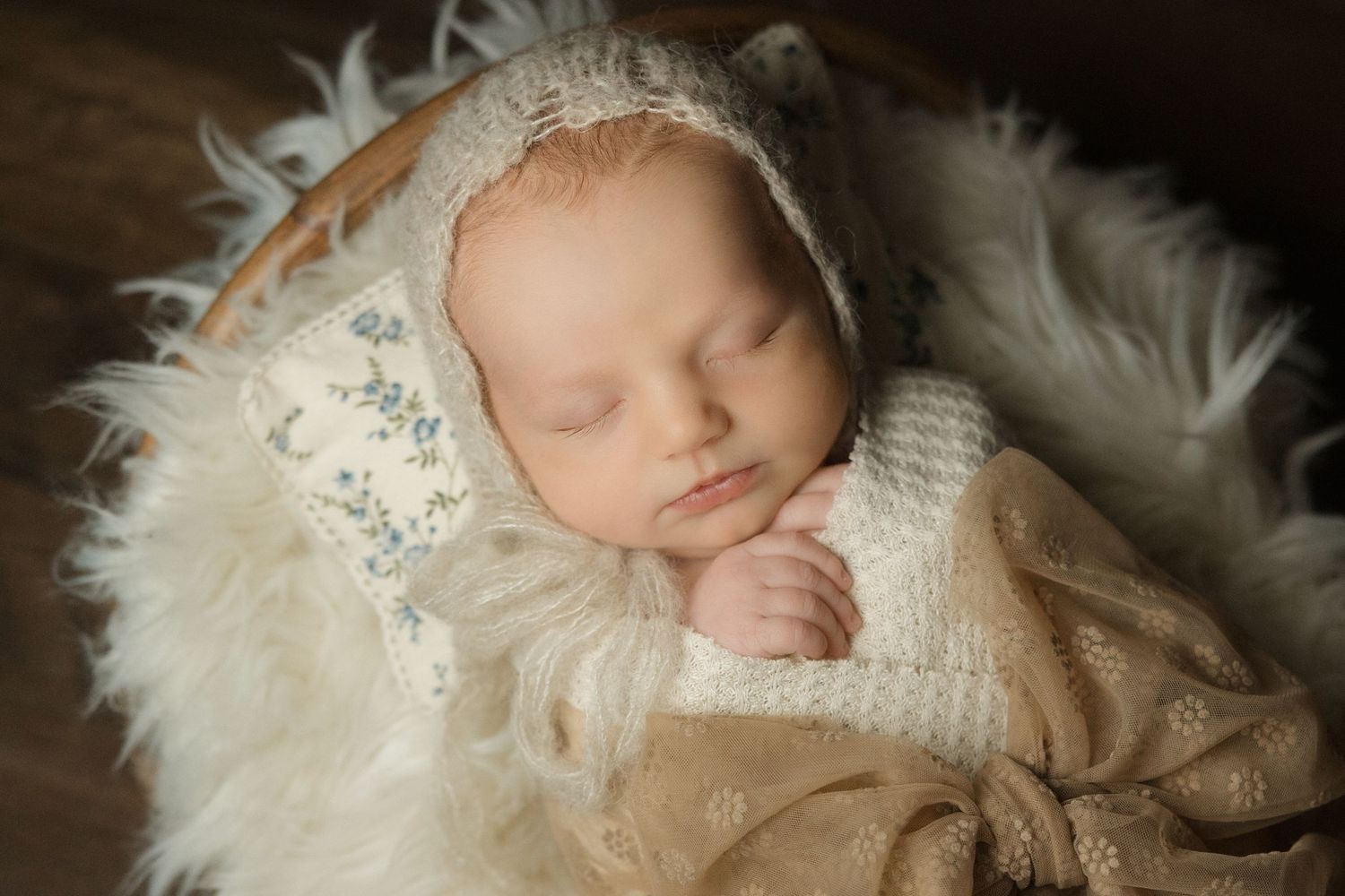 Peaceful newborn baby sleeping in a basket with fluffy white fur, wrapped in cream knit blanket and bonnet.