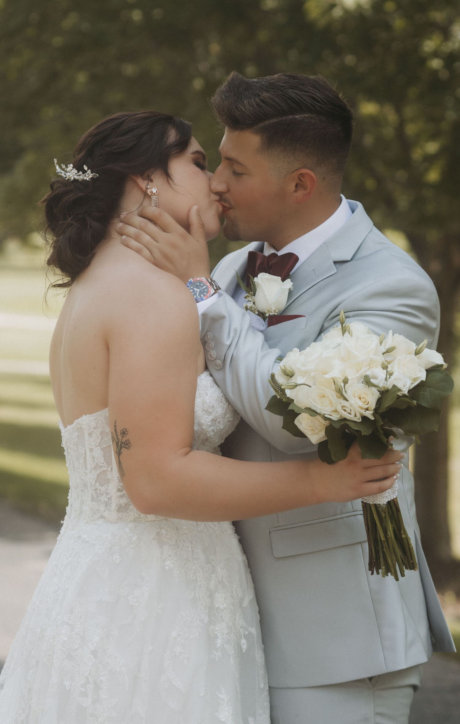 Wedding couple sharing romantic kiss while holding white rose bouquet outdoors.