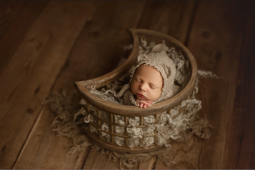 Newborn baby sleeping in a crescent moon-shaped basket with a knit bonnet on a rustic wood floor.