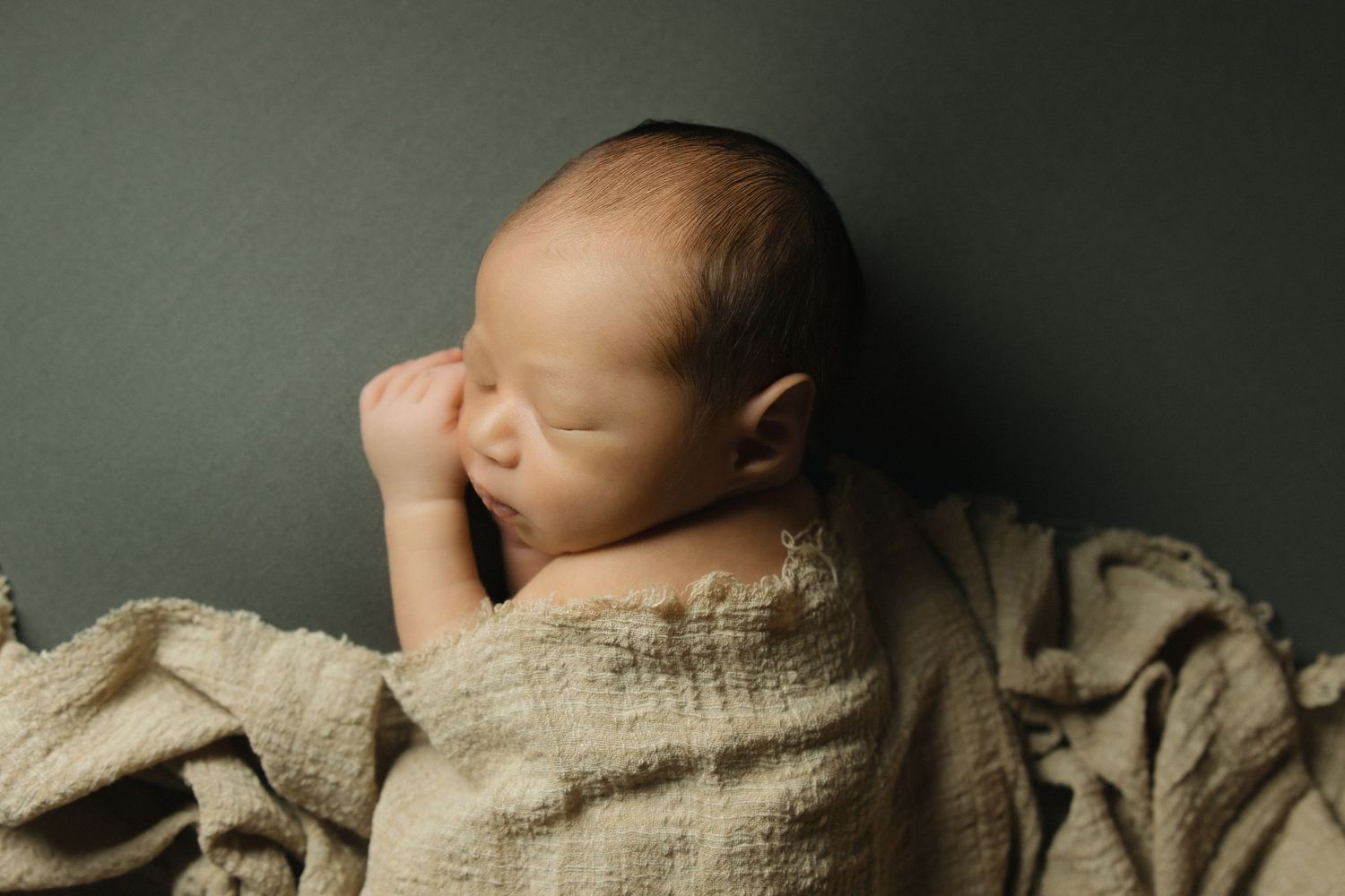 Peaceful newborn baby sleeping wrapped in a soft beige muslin cloth against a muted green backdrop.