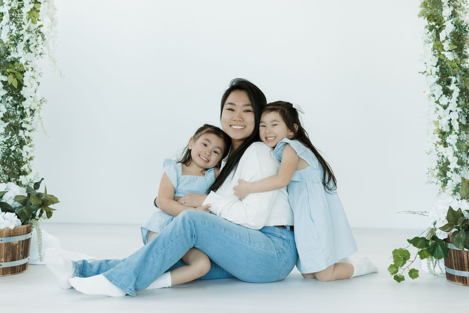 A smiling Asian woman sits on the floor with two young girls in matching blue outfits, posing for a family portrait.