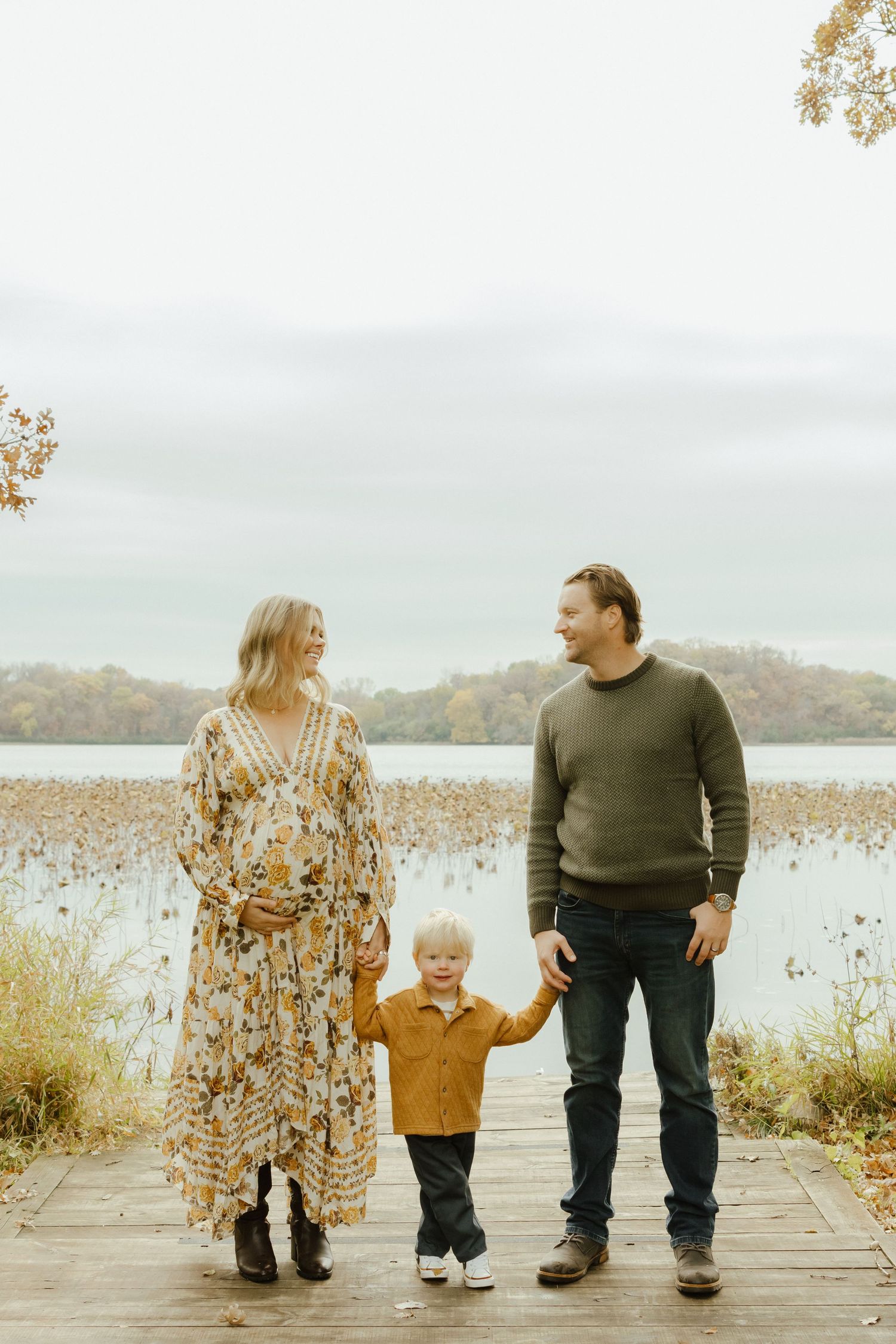 Pregnant woman in floral dress and man in green sweater hold toddler boy's hands on autumn lakeside dock.