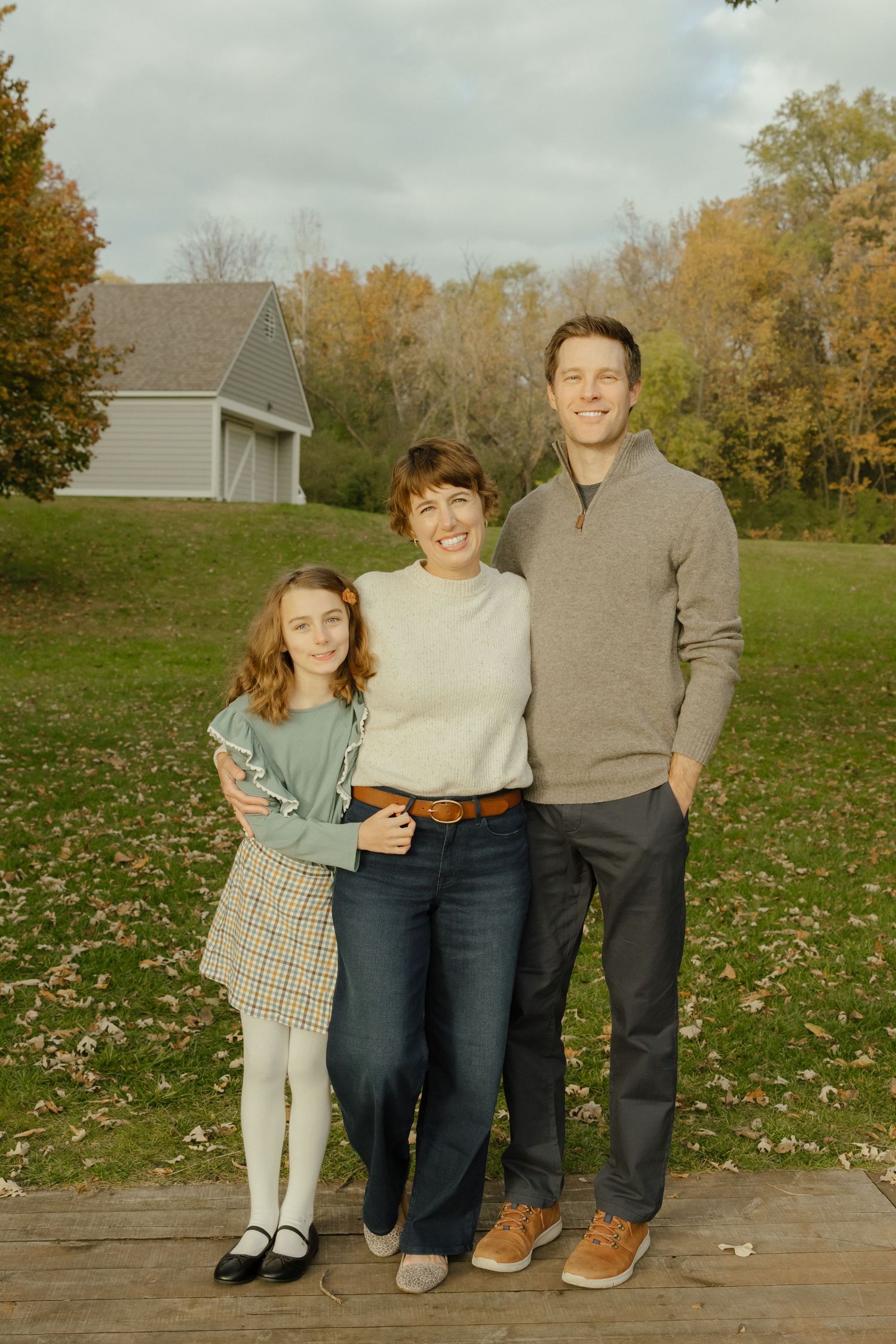Family of three smiling outdoors in autumn, standing on a wooden deck with colorful fall trees and a barn in background.